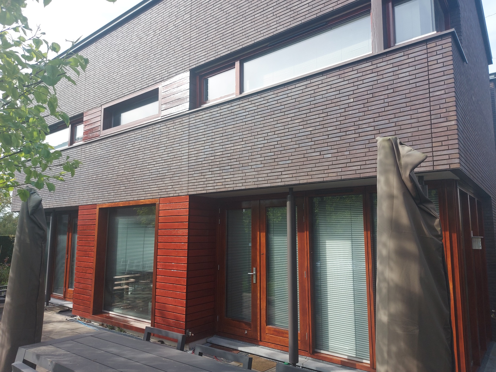 Nowoczesny dom z elewacją z cegły i drewnianymi elementami. Widoczne okna z żaluzjami i częściowo zasłonięty parasol ogrodowy. Ujęcie z dołu, podkreślające architekturę budynku.