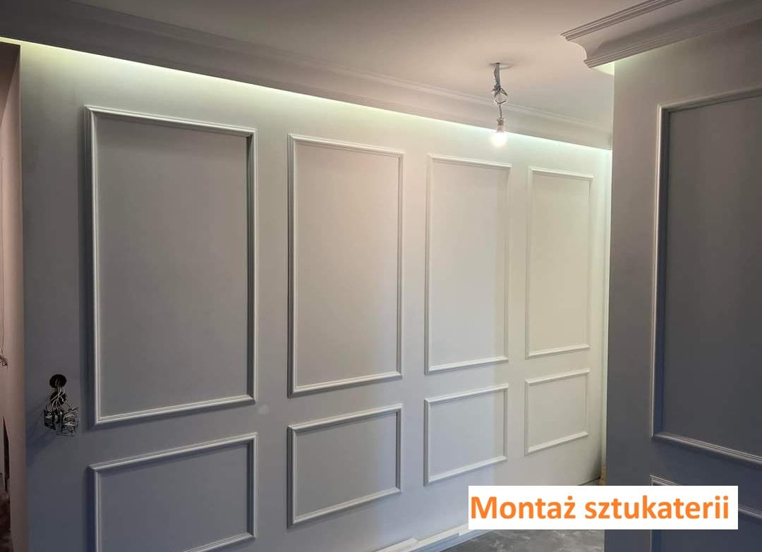 Ściana z zamontowaną białą sztukaterią w formie paneli, oświetlona od góry. Widoczny fragment sufitu z lampą. Dekoracyjne wykończenie wnętrza.