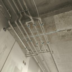Artem Bielikov - Szare rury instalacji wodnej i elektrycznej pod betonowym sufitem, mocowane metalowymi uchwytami. Widoczne napisy na rurach i fragment ściany w trakcie wykończenia.