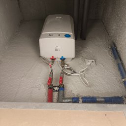 Artem Bielikov - Biały, wiszący bojler elektryczny marki Fox, z podłączonymi rurami (czerwona i niebieska) oraz kablem zasilającym, zamontowany w pomieszczeniu z szorstkimi ścianami.