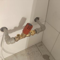 Artem Bielikov - Zamontowany zawór grzejnikowy z czerwoną głowicą termostatyczną, połączony z rurami wychodzącymi ze ściany wyłożonej białymi płytkami. Widoczne elementy mosiężne.