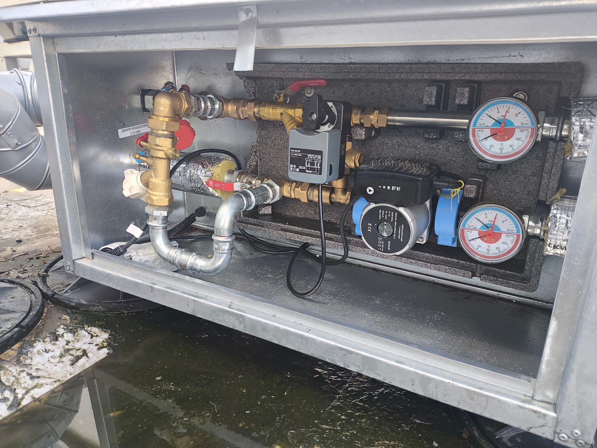 Węzeł cieplny w metalowej obudowie z widocznymi zaworami, pompami obiegowymi i manometrami. Instalacja w trakcie montażu, widoczne elementy izolacji.
