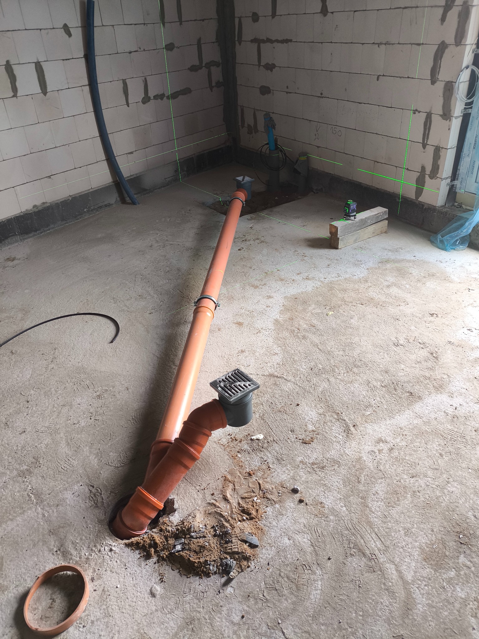 Pomarańczowe rury kanalizacyjne z kratką odpływową w surowym wnętrzu, widoczne ślady po wierceniu w betonie, poziomica laserowa wyznacza linie na ścianach z betonu komórkowego.