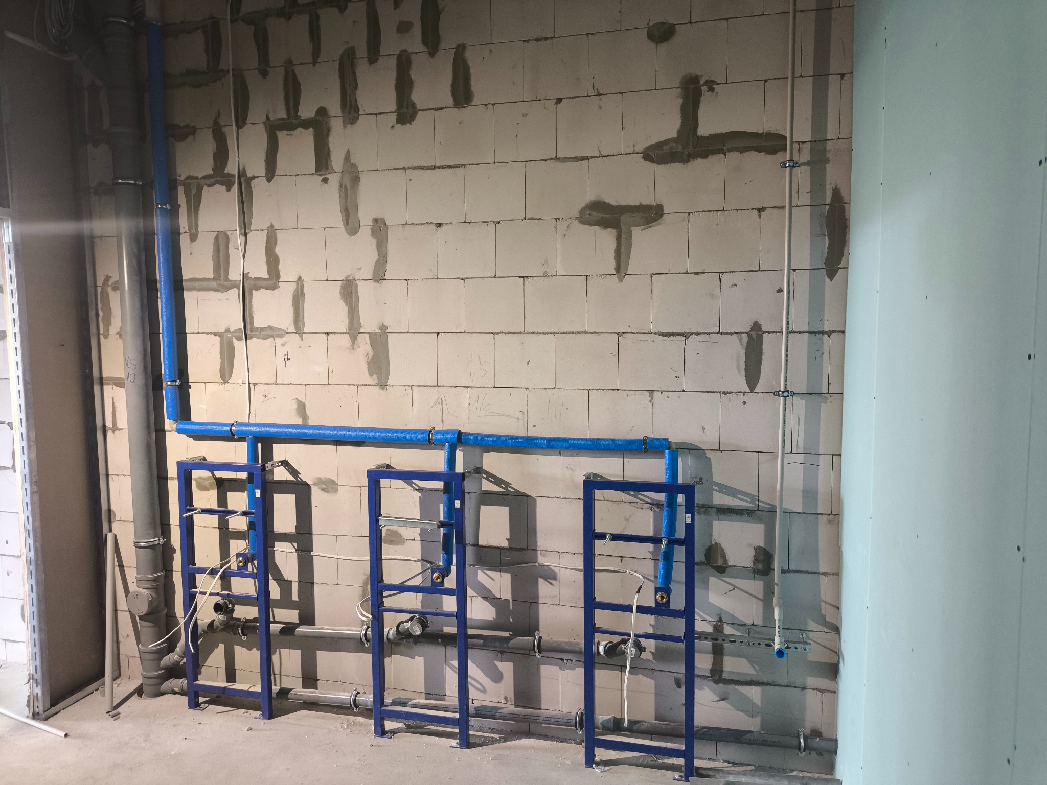 Niebieskie rury instalacji wodnej zamocowane na metalowych stelażach przy surowej ścianie z betonu komórkowego. Widoczne szare rury kanalizacyjne i piony instalacyjne.