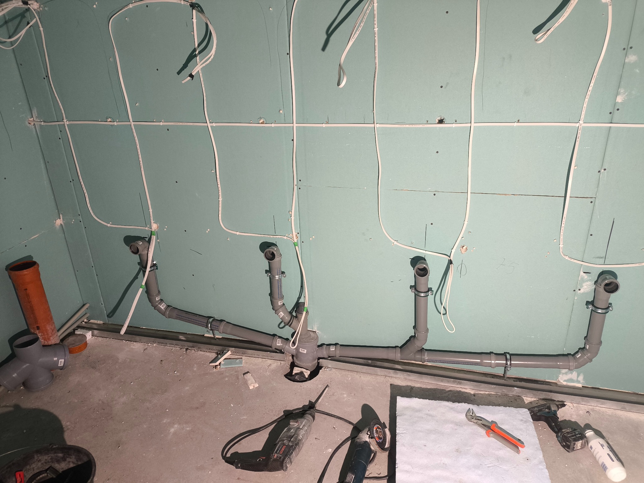 Szare rury kanalizacyjne zamontowane na ścianie z płyt gipsowo-kartonowych, widoczne narzędzia i kable. Instalacja w trakcie montażu, surowy stan wykończenia.