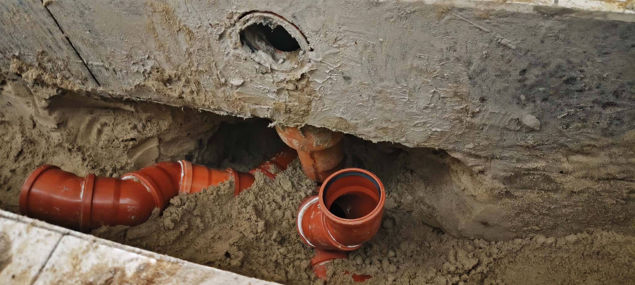 Pomarańczowe rury kanalizacyjne ułożone w wykopie z piasku, widoczne otwory w ścianie. Instalacja w trakcie prac, surowe wykończenie.