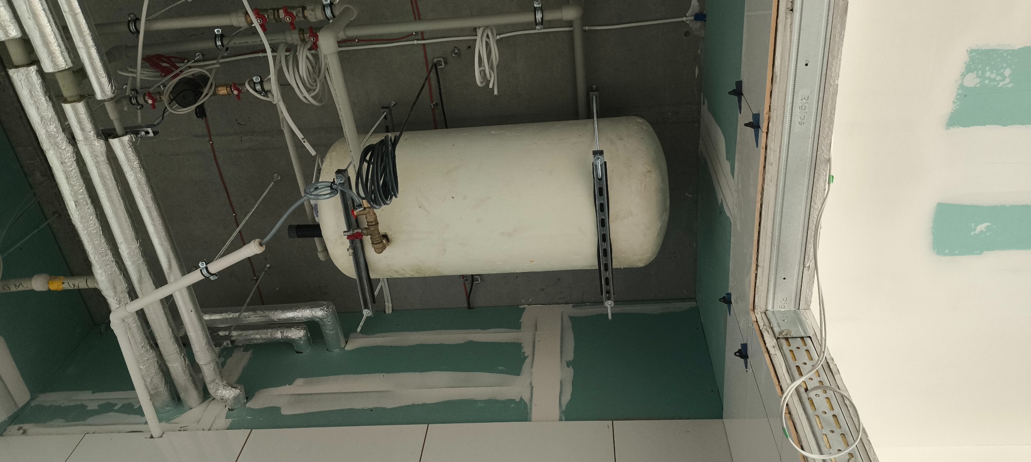 Montaż poziomego bojlera w surowym wnętrzu. Widoczne rury, kable i elementy konstrukcyjne ściany. Ujęcie z dołu, podkreślające instalację w trakcie realizacji.