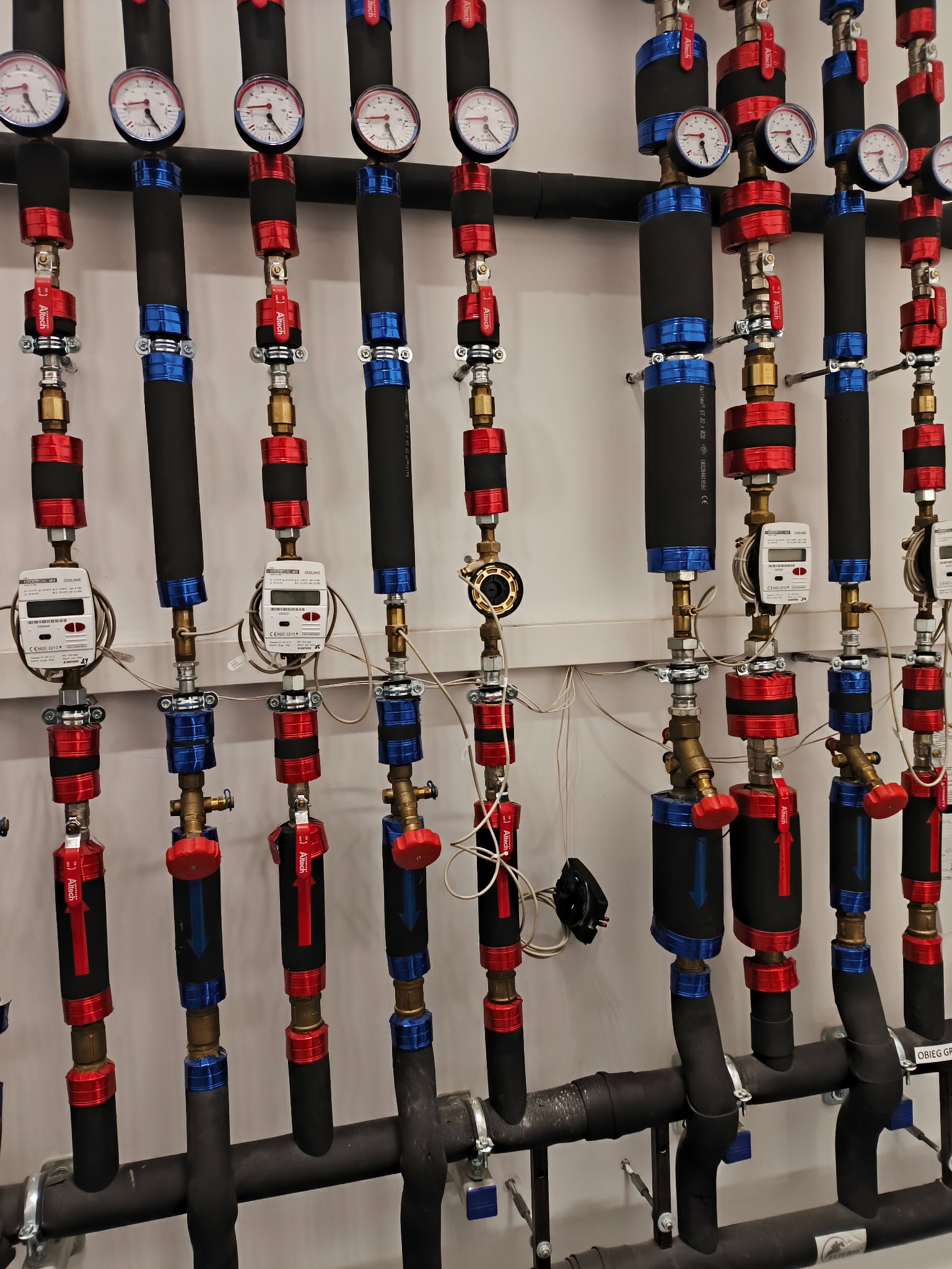 Rozbudowana instalacja grzewcza z czarnymi rurami, czerwonymi i niebieskimi elementami, manometrami i licznikami ciepła na białej ścianie. Widoczne zawory i izolacja.