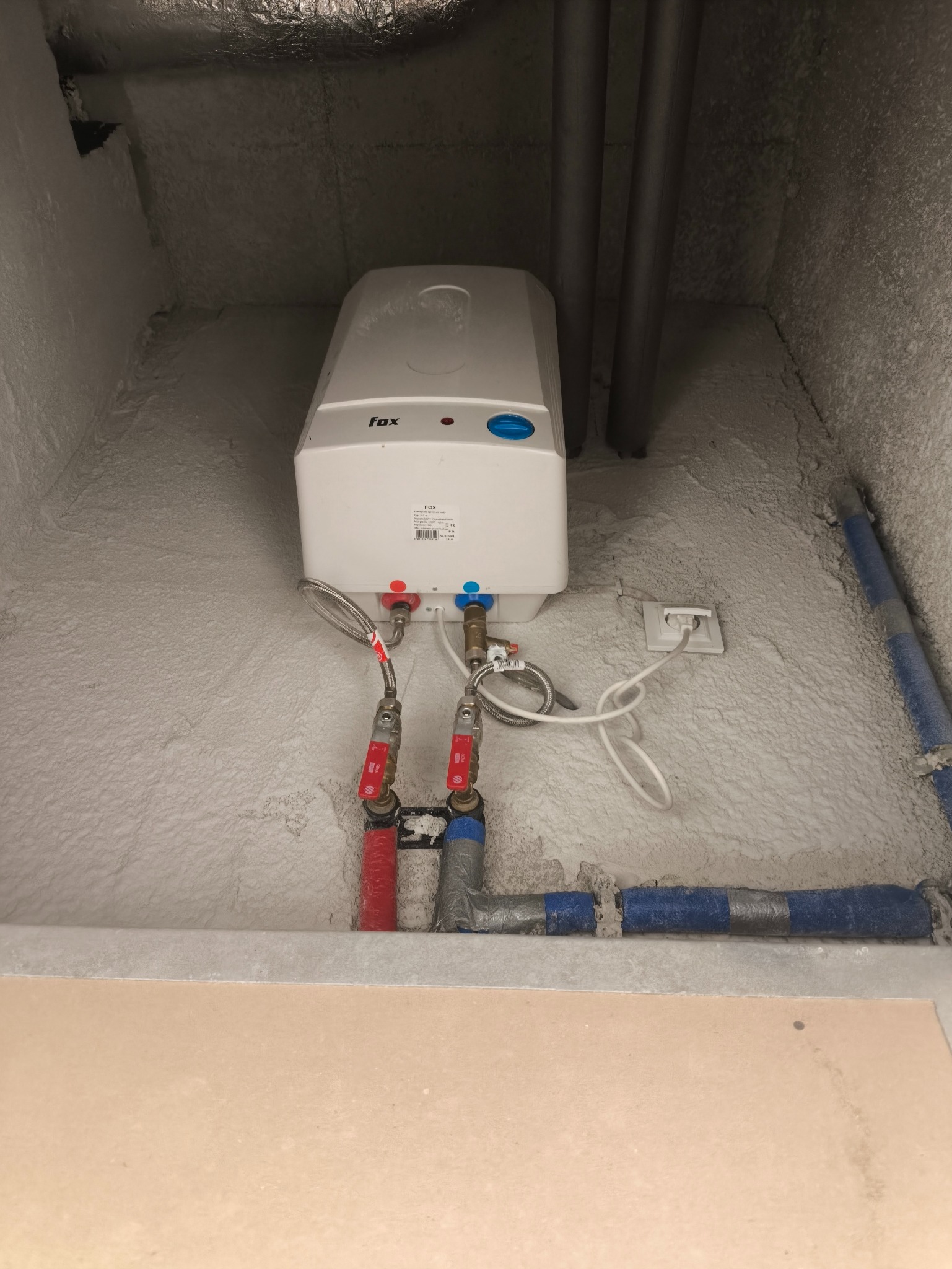 Biały, wiszący bojler elektryczny marki Fox, z podłączonymi rurami (czerwona i niebieska) oraz kablem zasilającym, zamontowany w pomieszczeniu z szorstkimi ścianami.