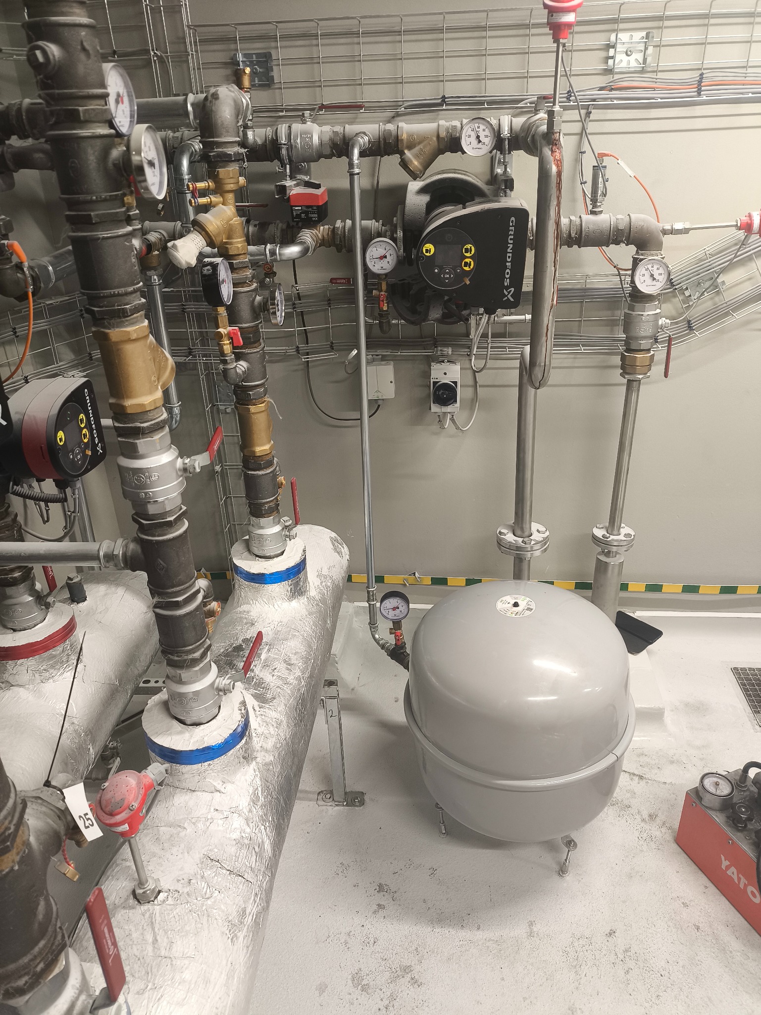 Złożona instalacja grzewcza z rurami, zaworami, manometrami i pompą obiegową Grundfos w kotłowni. Widoczne izolowane rury i zbiornik wyrównawczy.