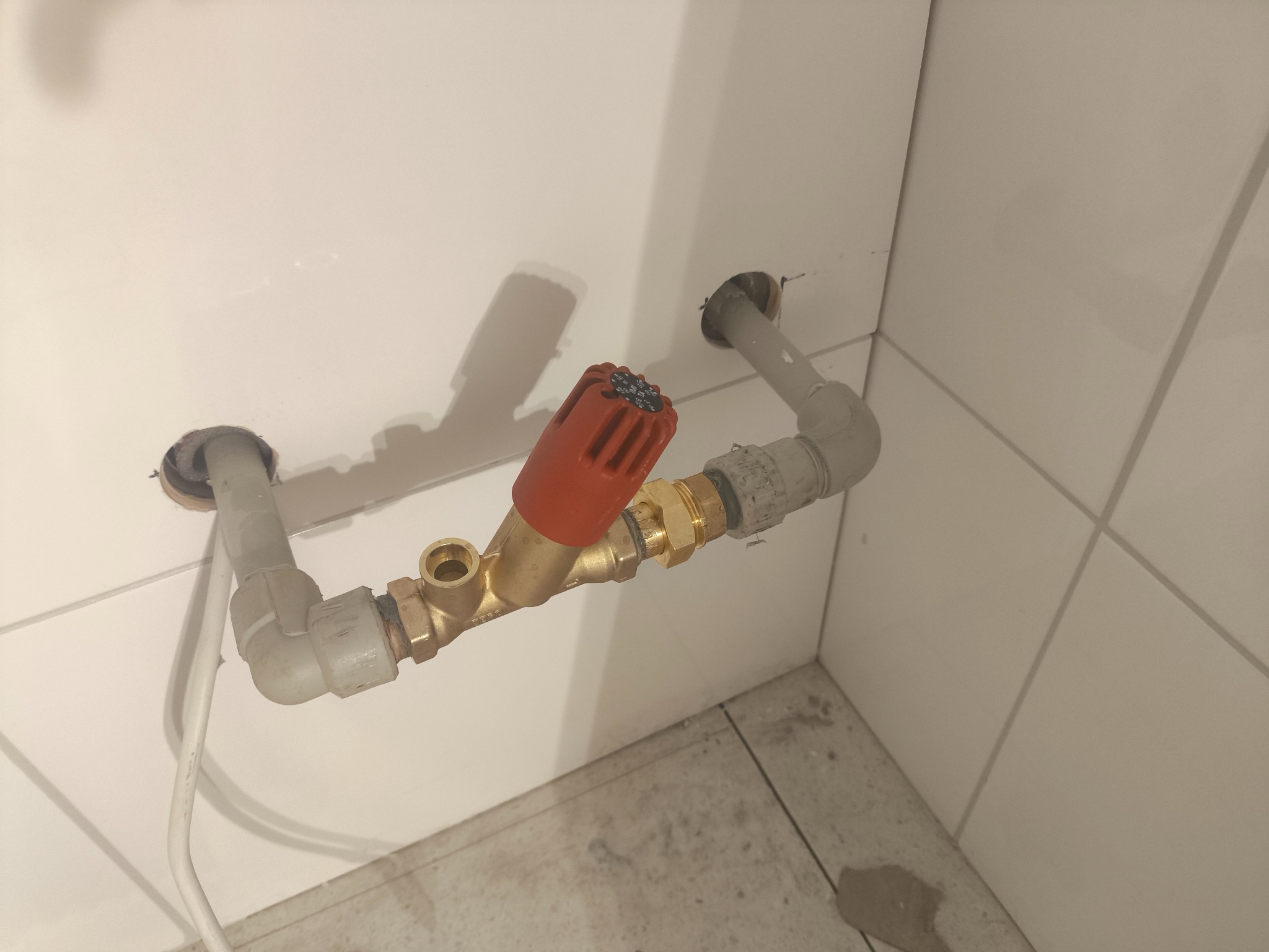 Zamontowany zawór grzejnikowy z czerwoną głowicą termostatyczną, połączony z rurami wychodzącymi ze ściany wyłożonej białymi płytkami. Widoczne elementy mosiężne.