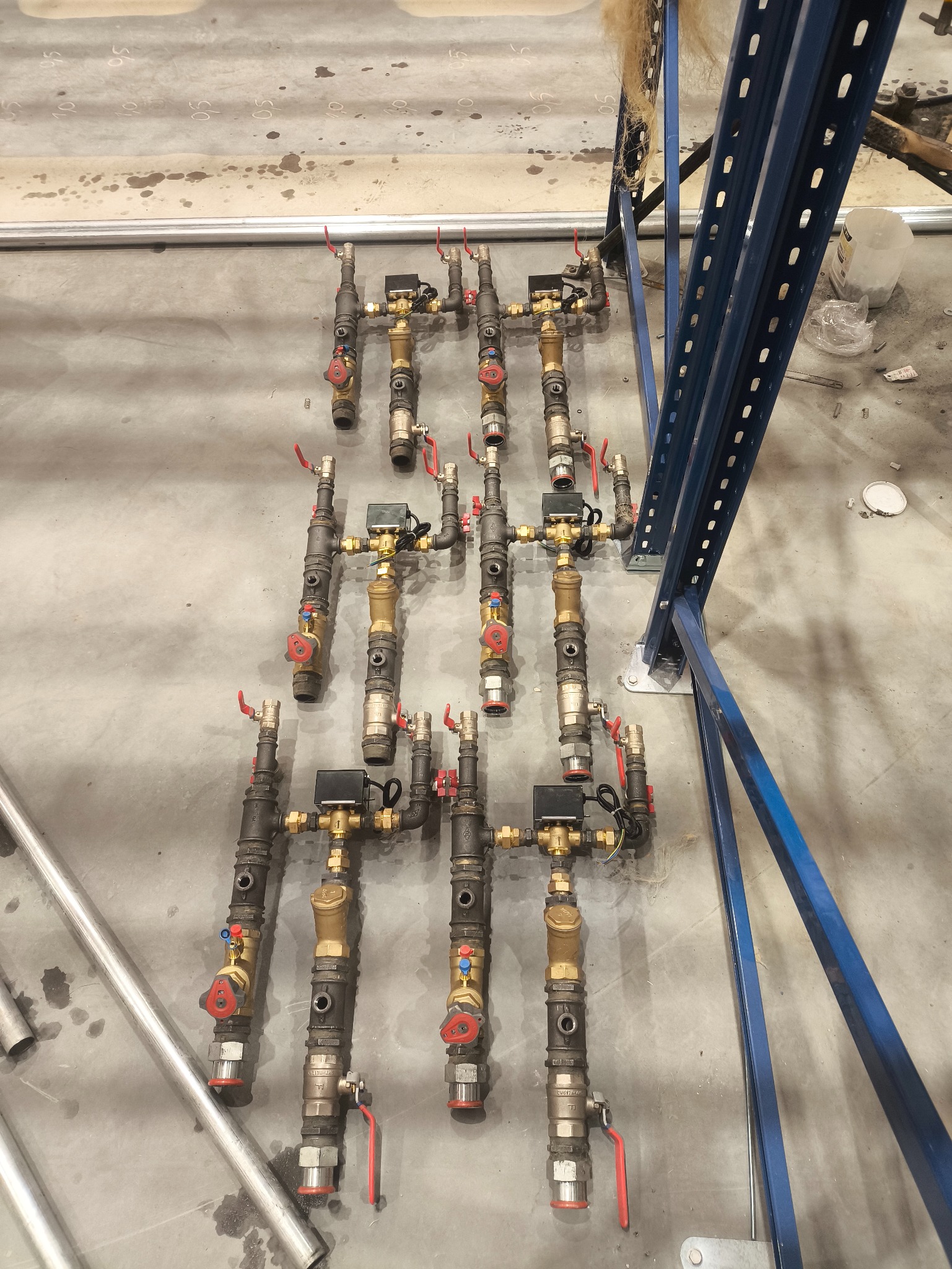 Rozdzielacze instalacji wodnej z zaworami i licznikami, ułożone rzędami na szarym betonowym podłożu. Widoczne elementy metalowej konstrukcji regału.