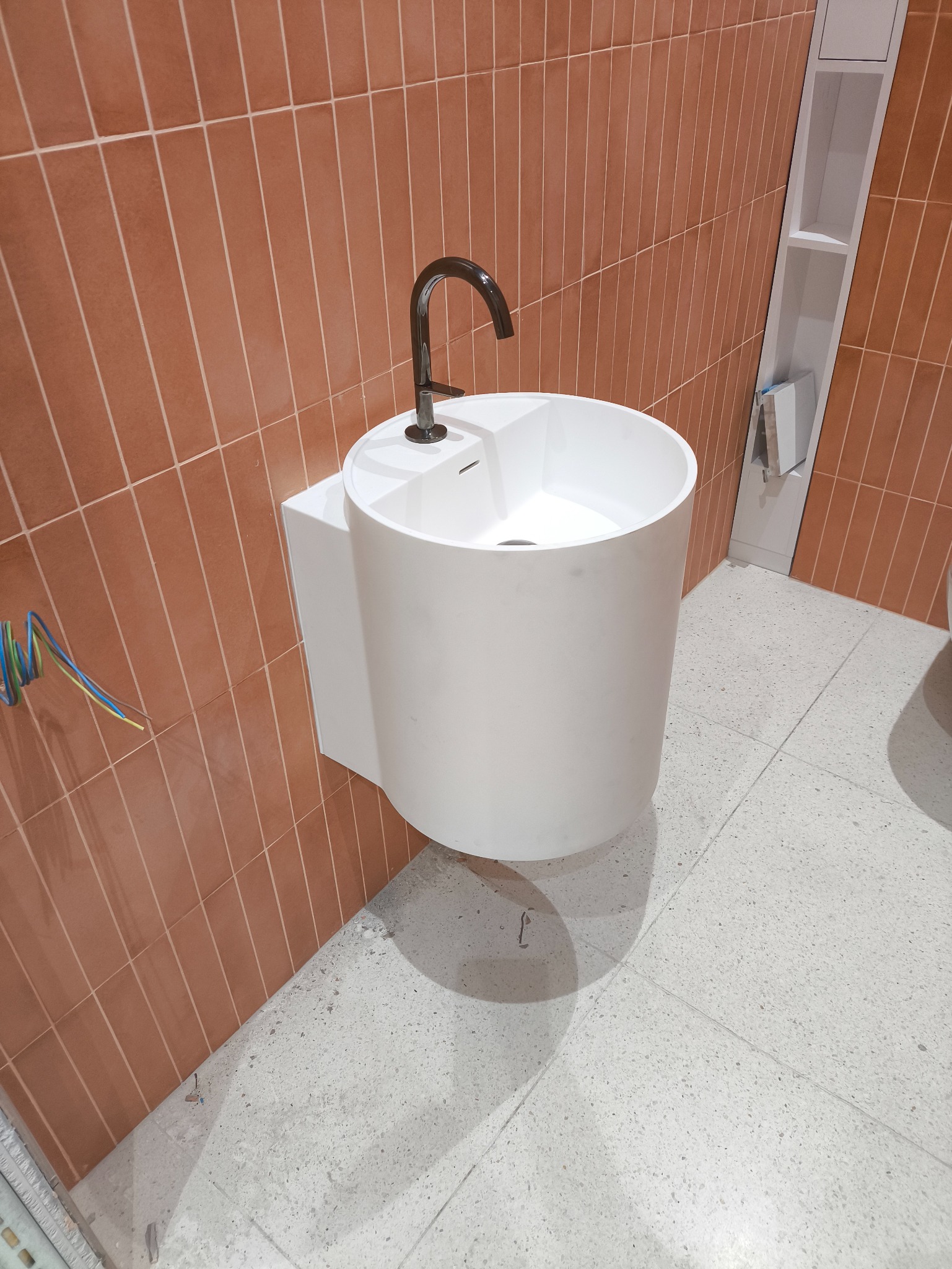 Nowoczesna, biała umywalka wolnostojąca z czarną armaturą na tle ściany z terakotowych płytek i jasnej podłogi z lastryko. Widoczne przewody instalacyjne.