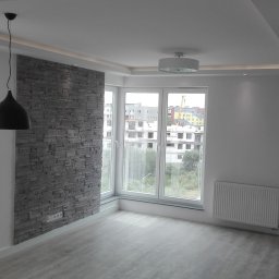 DUM - BUD ANDRZEJ DUMANOWSKI - Jasne wnętrze po remoncie: ściana z dekoracyjnej szarej cegły, duże okna z widokiem na miasto, szara podłoga imitująca drewno, nowoczesne oświetlenie sufitowe.