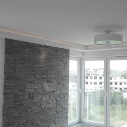 DUM - BUD ANDRZEJ DUMANOWSKI - Pokój po wykończeniu: ściana z szarej okładziny kamiennej, narożne okno z widokiem na zewnątrz, nowoczesna lampa sufitowa i jasna podłoga. Minimalistyczny design.