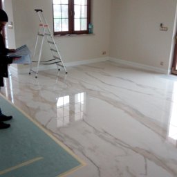 DUM - BUD ANDRZEJ DUMANOWSKI - Położona, błyszcząca, marmurowa podłoga w jasnym pomieszczeniu. Widoczna drabina, okno i fragment osoby z planami. Podłoga zabezpieczona folią malarską.