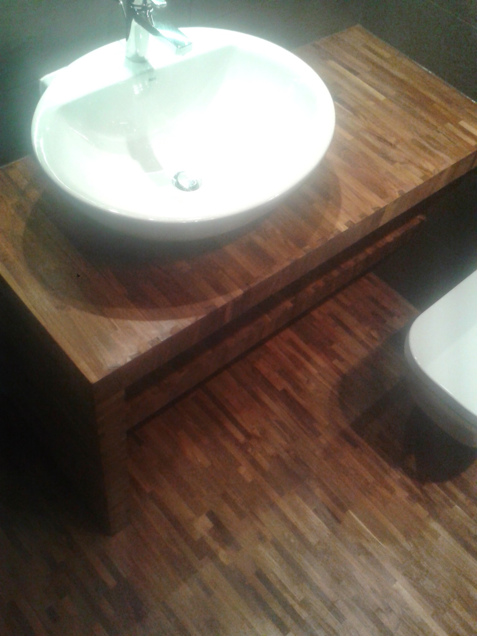 Nowoczesna łazienka z drewnianym blatem pod umywalkę i parkietem w ciepłych odcieniach brązu. Biała umywalka z chromowaną baterią.