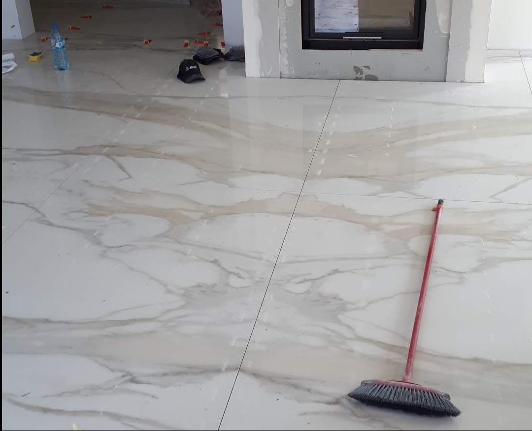 Położona, błyszcząca podłoga z płytek imitujących marmur, z widoczną szczotką do zamiatania i akcesoriami glazurniczymi w tle. Widok na fragment kominka.