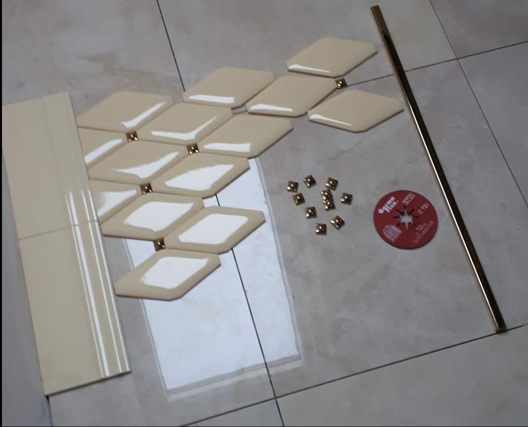 Beżowe, romboidalne płytki ułożone wzorem, złote łączniki, listwa wykończeniowa i tarcza do cięcia płytek na jasnej, błyszczącej powierzchni.