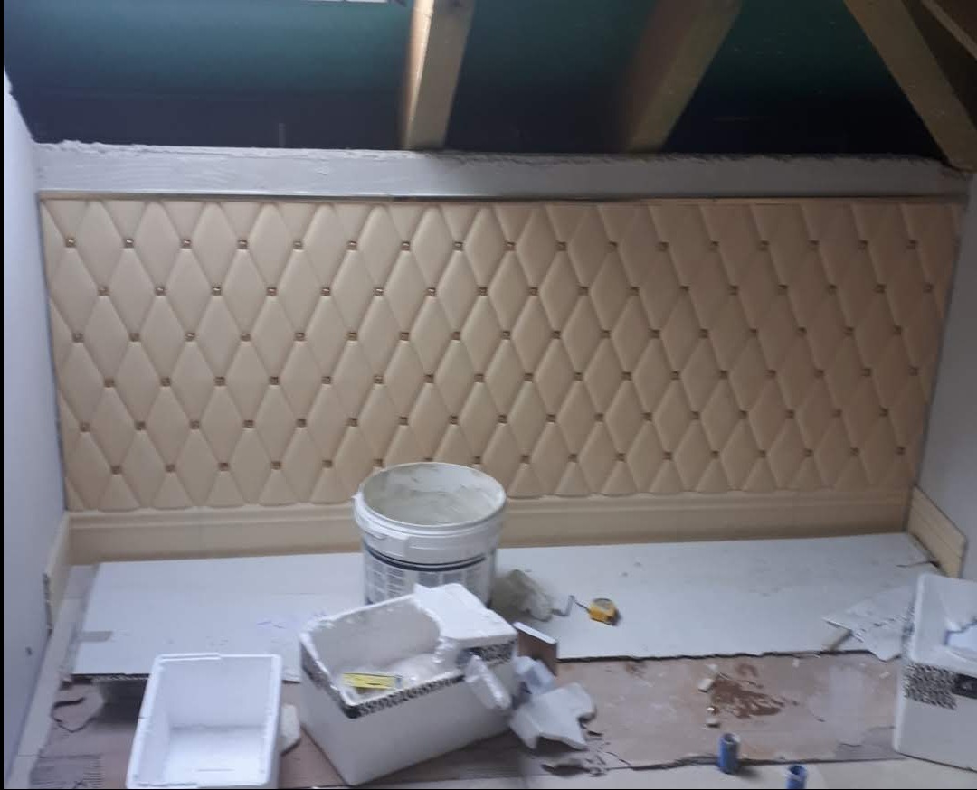 Beżowa, pikowana ściana w skosie poddasza z dekoracyjnymi guzikami. Widoczne narzędzia i materiały budowlane. Prace wykończeniowe w trakcie realizacji.