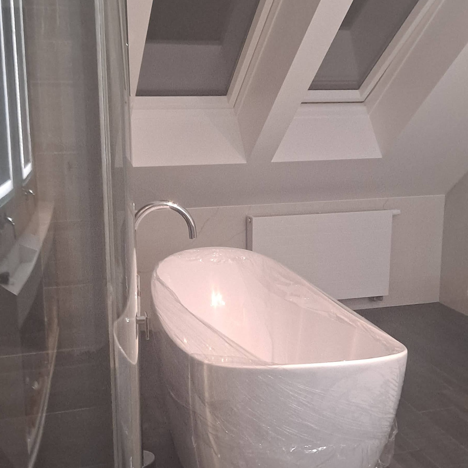 Nowoczesna, biała wanna wolnostojąca, jeszcze w folii ochronnej, w jasnej łazience na poddaszu z oknami dachowymi. Chromowana bateria. Minimalistyczny grzejnik.
