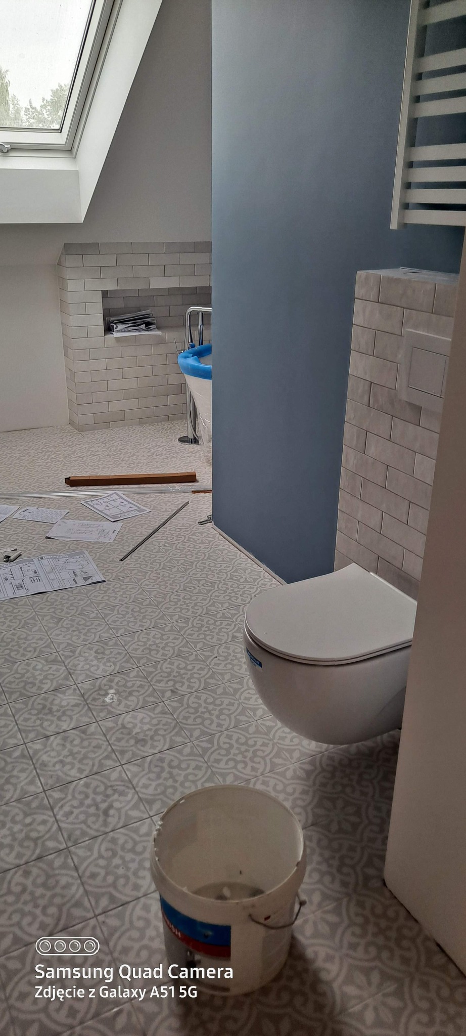 Ukończona łazienka na poddaszu z wzorzystą podłogą, białą armaturą i niebieską ścianą. Widoczne plany i narzędzia na podłodze, co sugeruje świeży remont.