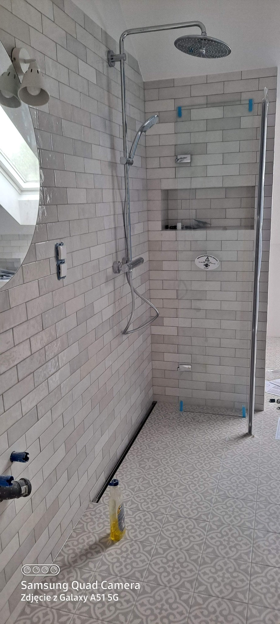 Wykończona łazienka z kabiną prysznicową typu walk-in, jasnymi płytkami imitującymi cegłę i wzorzystą podłogą. Widoczny odpływ liniowy i bateria prysznicowa.