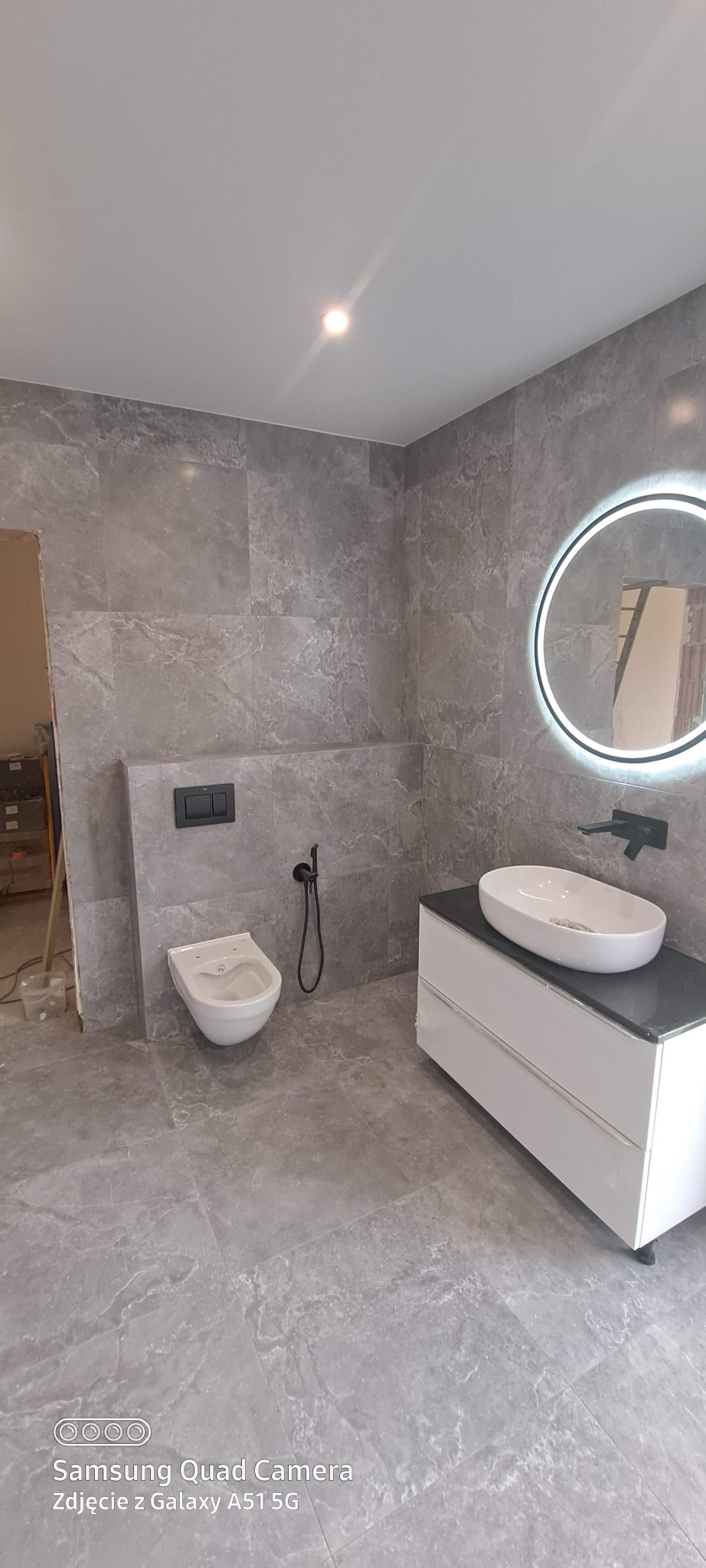 Wykończona łazienka w szarościach: marmurowe płytki, podwieszana toaleta z bidetką, biała szafka z umywalką i okrągłe lustro z podświetleniem LED. Minimalistyczny design.