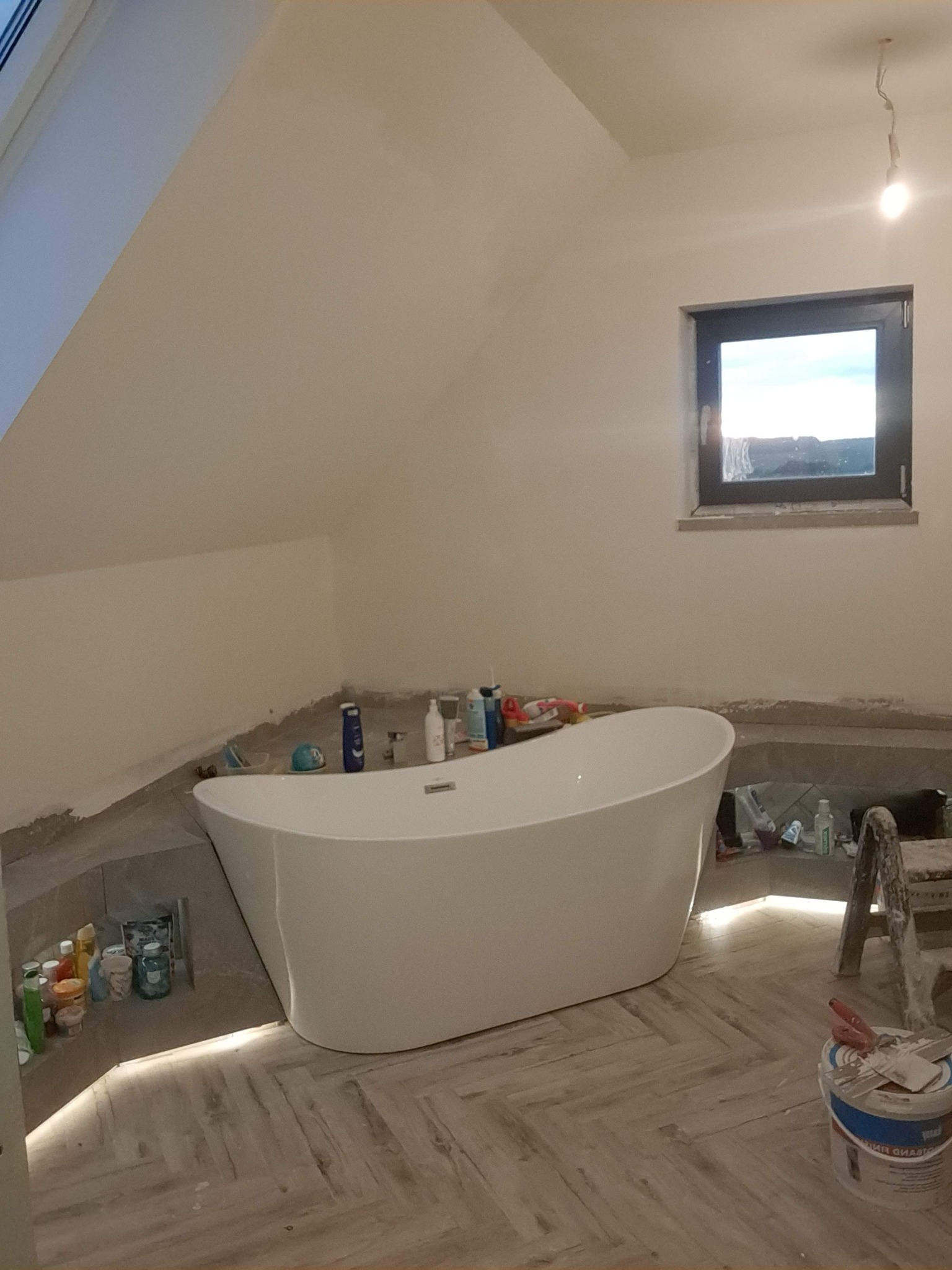 Biała wanna wolnostojąca w łazience na poddaszu z oknem. Ułożona podłoga w jodełkę, widoczne materiały budowlane i oświetlenie LED przy wannie. Etap prac wykończeniowych.