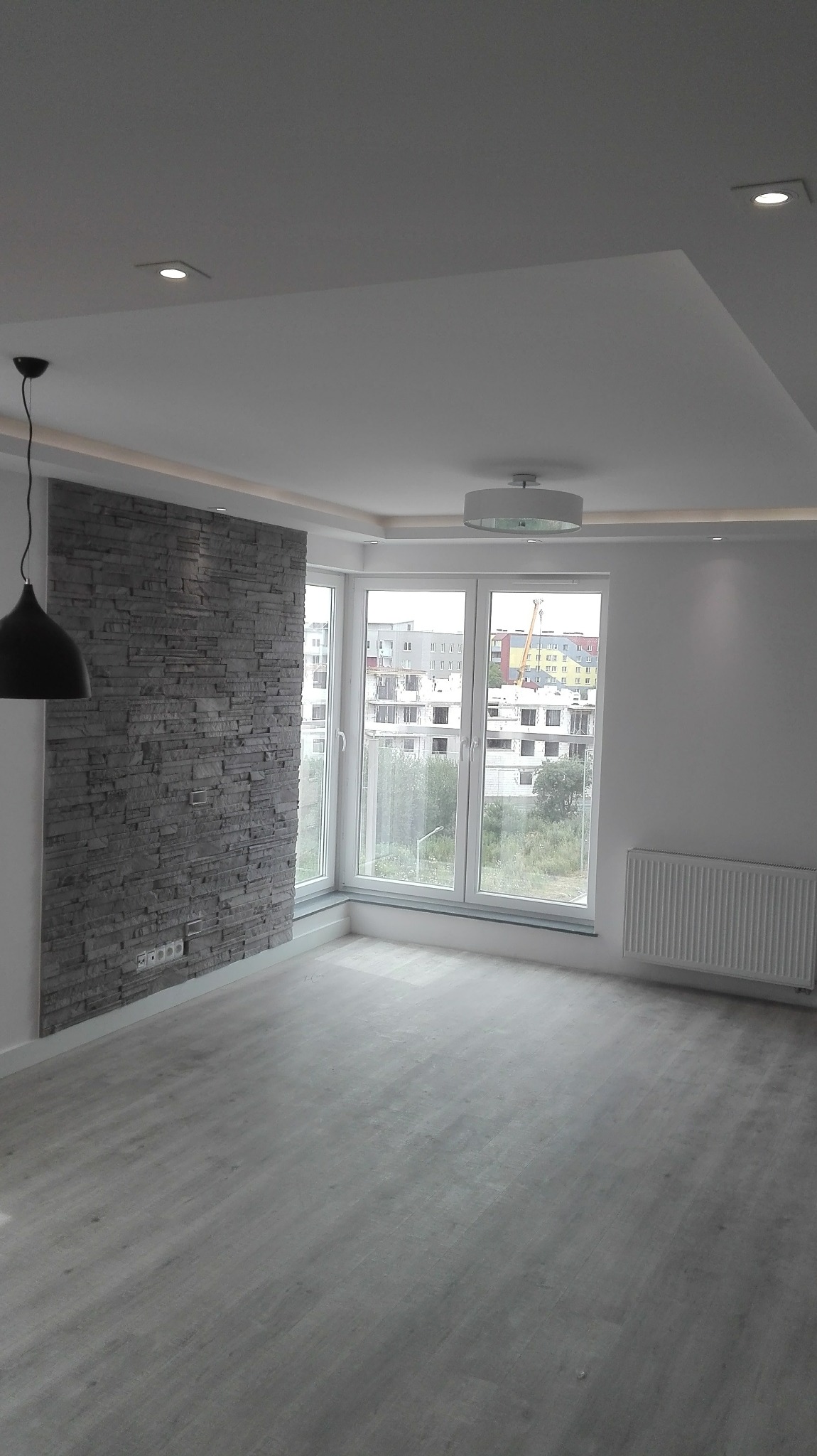 Jasne wnętrze po remoncie: ściana z dekoracyjnej szarej cegły, duże okna z widokiem na miasto, szara podłoga imitująca drewno, nowoczesne oświetlenie sufitowe.