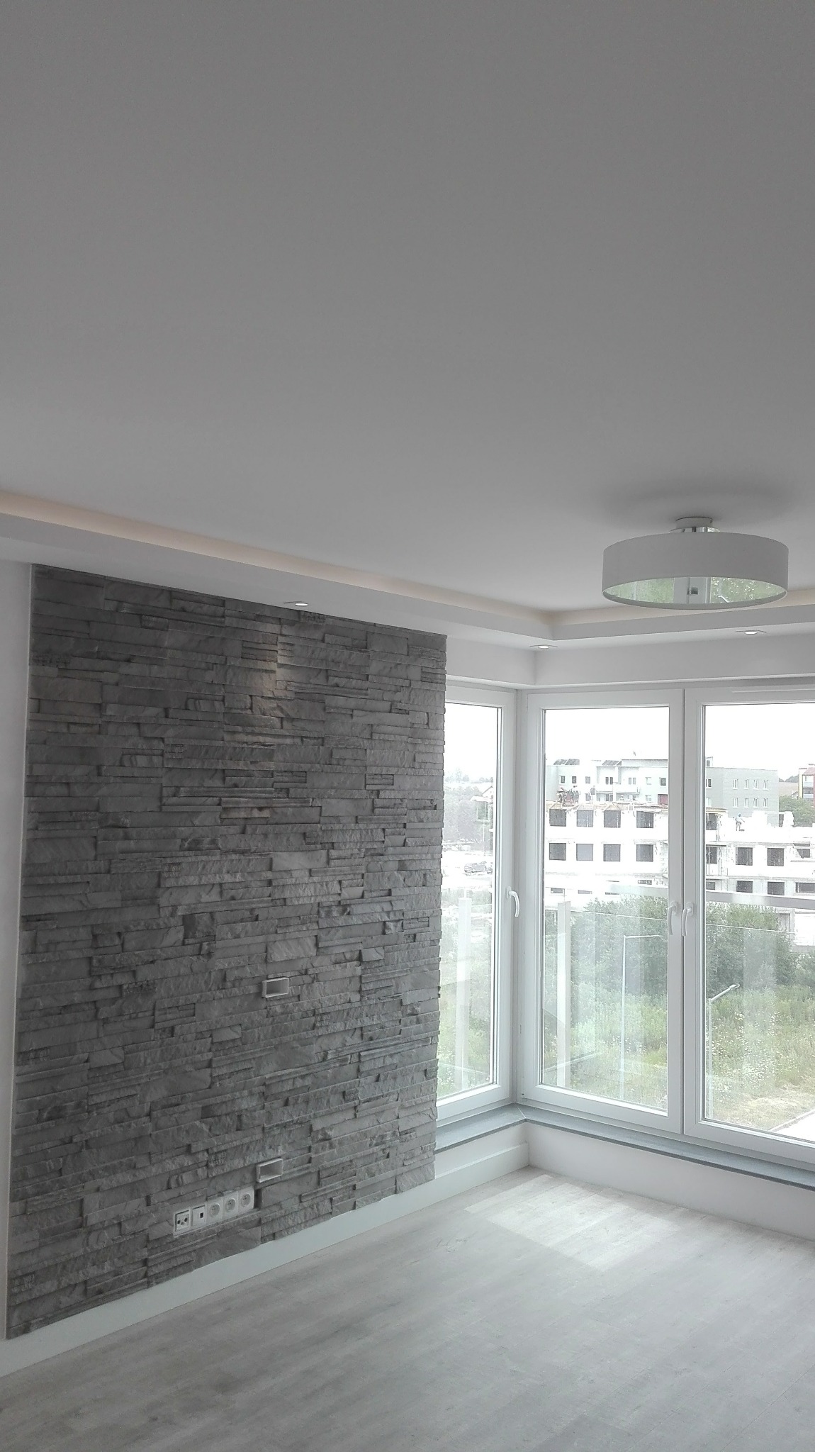 Pokój po wykończeniu: ściana z szarej okładziny kamiennej, narożne okno z widokiem na zewnątrz, nowoczesna lampa sufitowa i jasna podłoga. Minimalistyczny design.