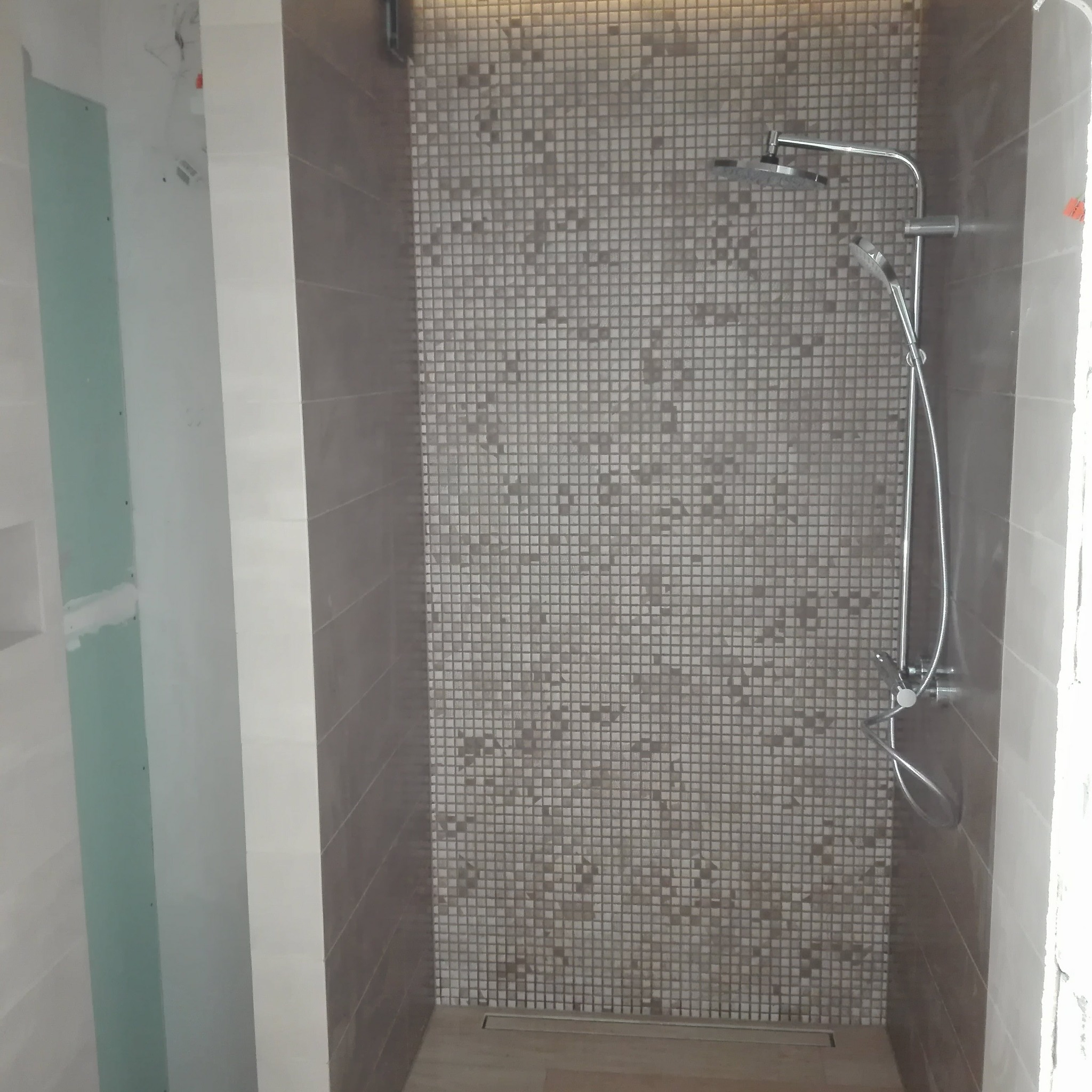 Kabina prysznicowa z mozaiką w odcieniach szarości i beżu. Widoczna armatura i odpływ liniowy. Częściowo wykończona ściana z płyt karton-gips.