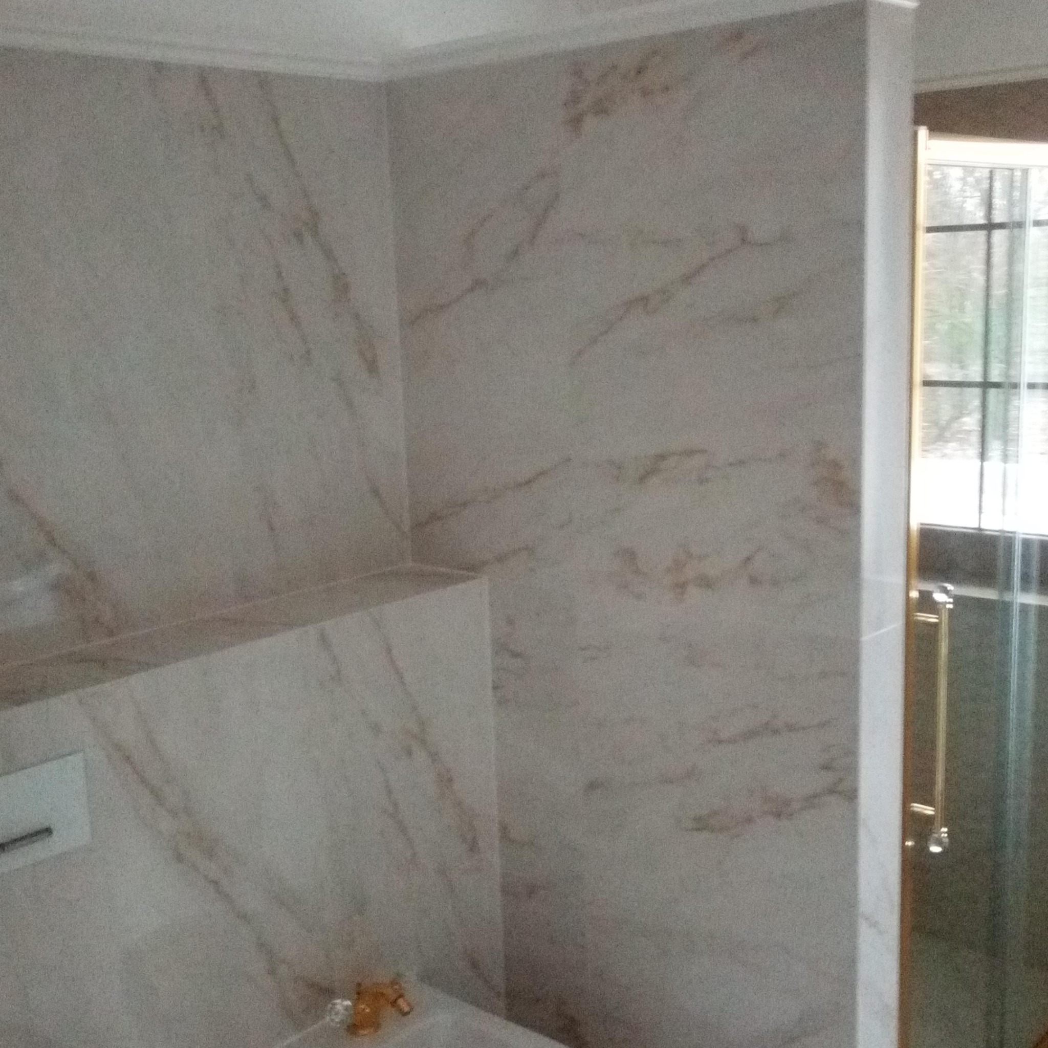 Wykończona łazienka z marmurowymi płytkami w beżowych odcieniach. Widoczny fragment kabiny prysznicowej z złotym uchwytem. Minimalistyczny styl, jasne wnętrze.