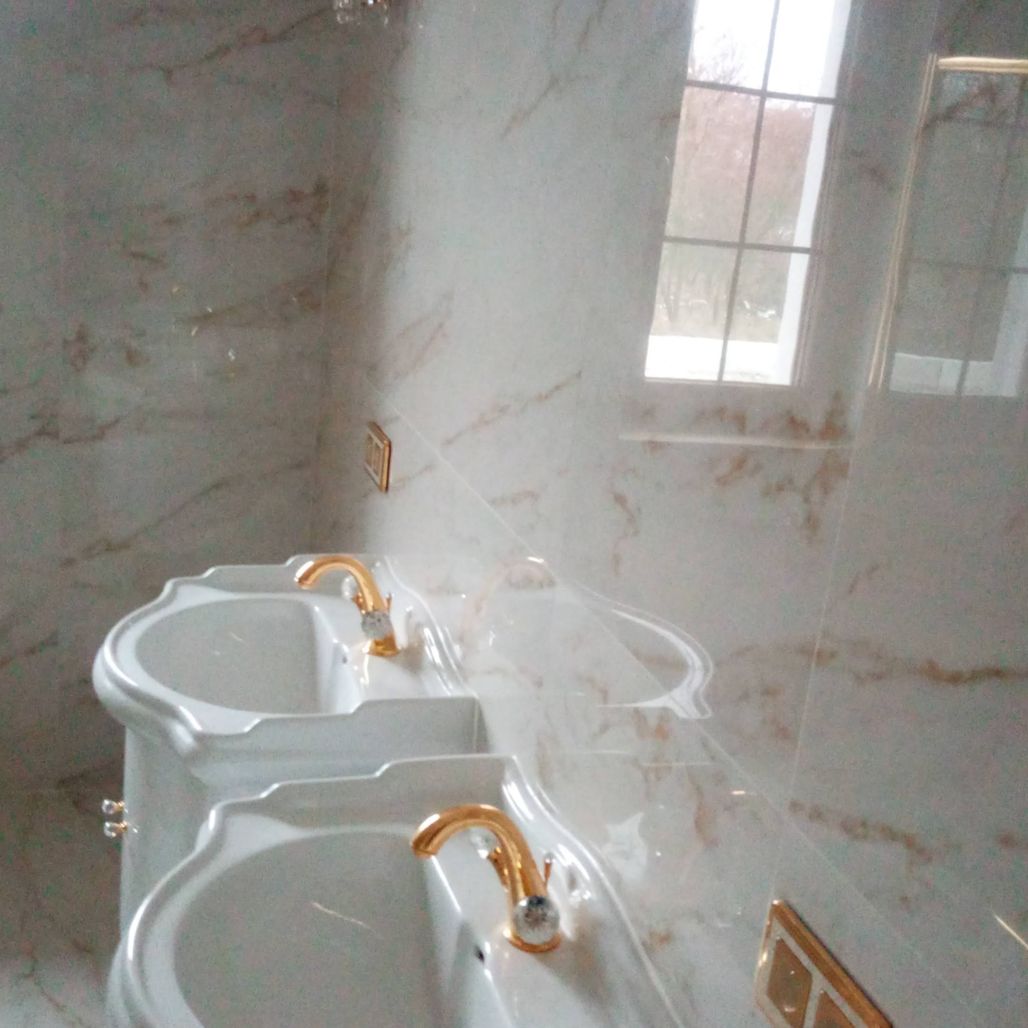 Elegancka łazienka z marmurowymi płytkami i podwójną umywalką z złotymi kranami. Widoczne okno z naturalnym światłem. Luksusowe wykończenie wnętrza.