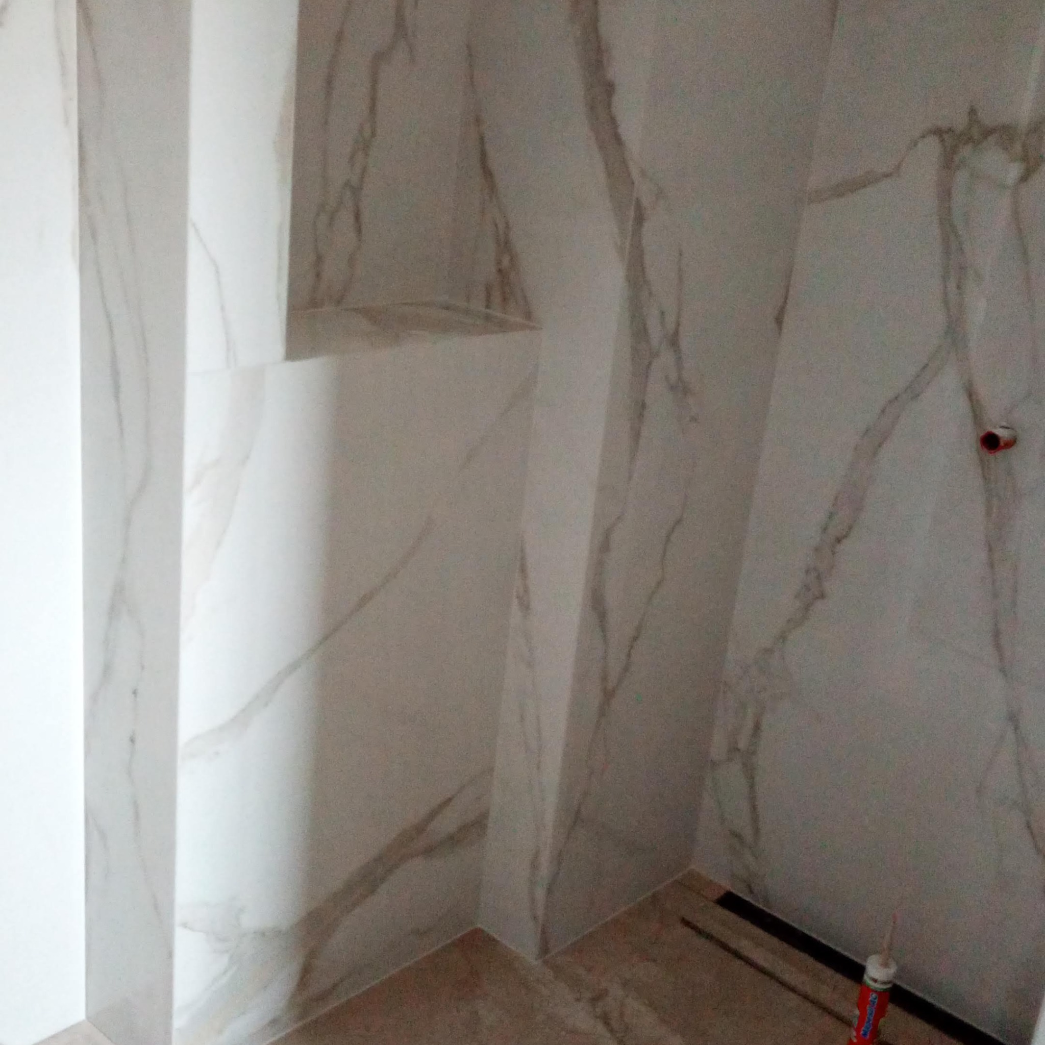 Wykończona ściana w łazience z płytkami imitującymi marmur, widoczny fragment podłogi i narożnik. U dołu leży tuba silikonu.