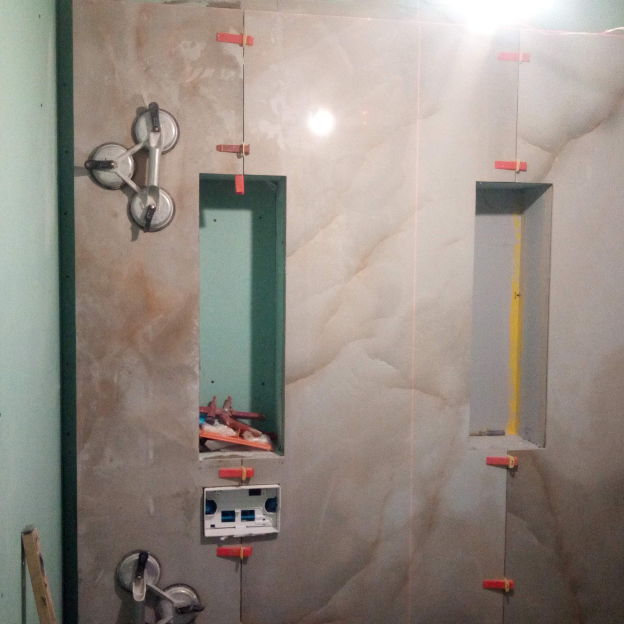 Montaż płytek w łazience z użyciem przyssawek i poziomowania. Widoczne wnęki w ścianie i puszka elektryczna. Laserowy wskaźnik wyznacza linię.