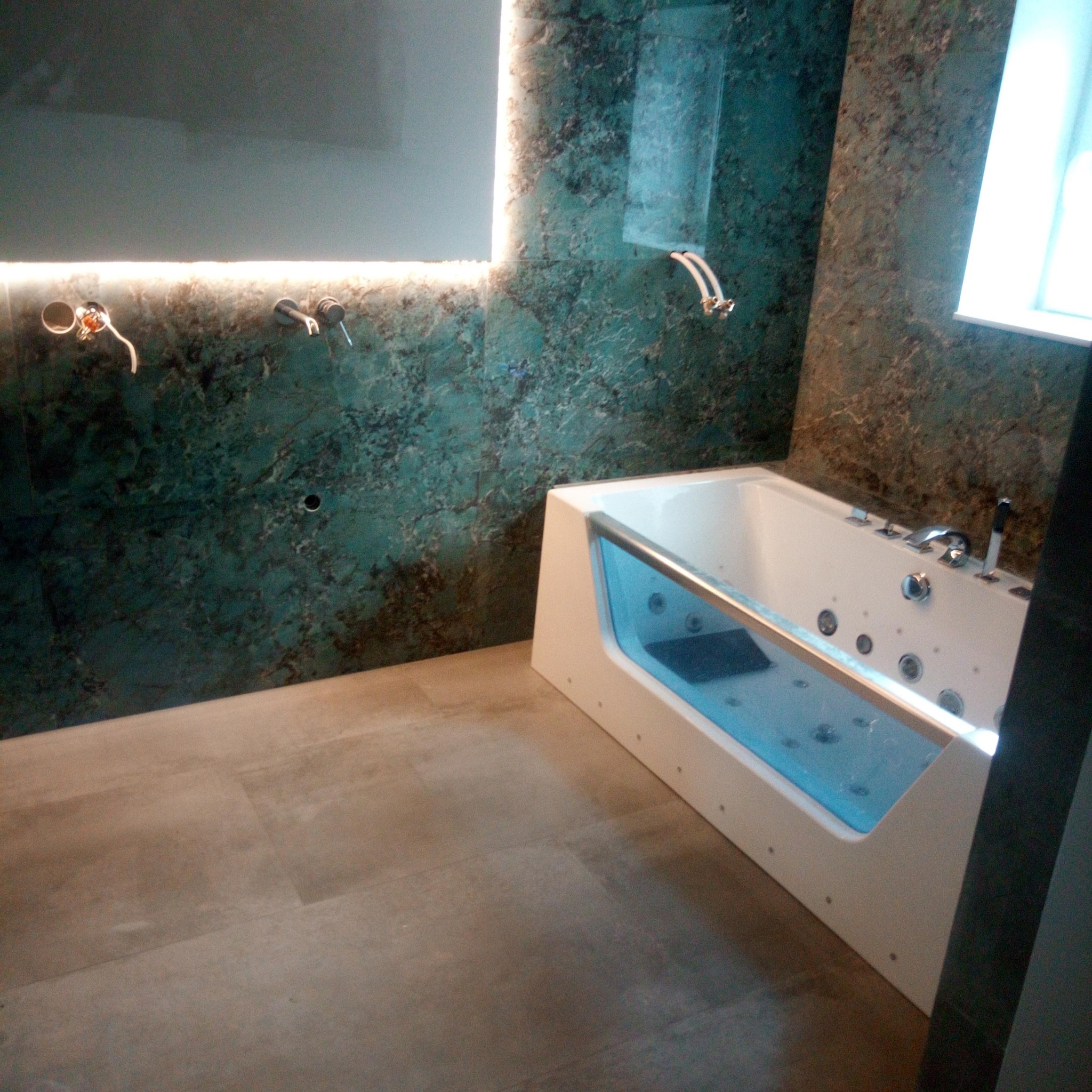 Nowoczesna łazienka z wanną z hydromasażem i podświetlanym lustrem. Ściany wykończone płytkami imitującymi kamień w odcieniach zieleni i brązu. Minimalistyczna armatura.