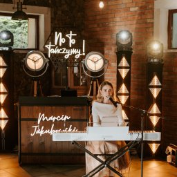 Agnieszka Osiecka - Wokalistka z mikrofonem śpiewa, grając na keyboardzie Casio. W tle ceglana ściana, neon 'No to tańczymy!' i oświetlenie sceniczne. Marcin Jakubowski.