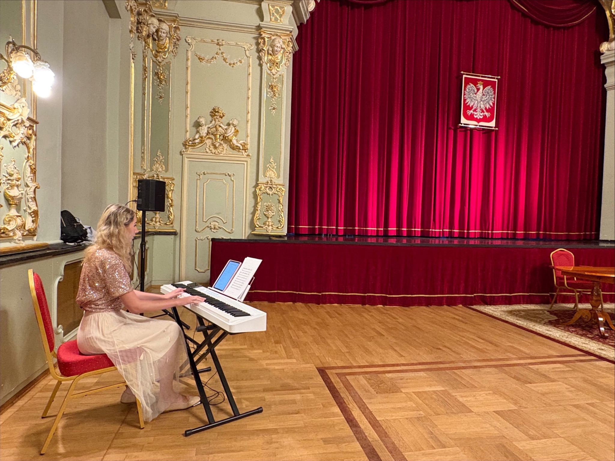Blondynka gra na keyboardzie w bogato zdobionej sali balowej z czerwonymi zasłonami i godłem Polski. Scena gotowa na występ artystyczny.