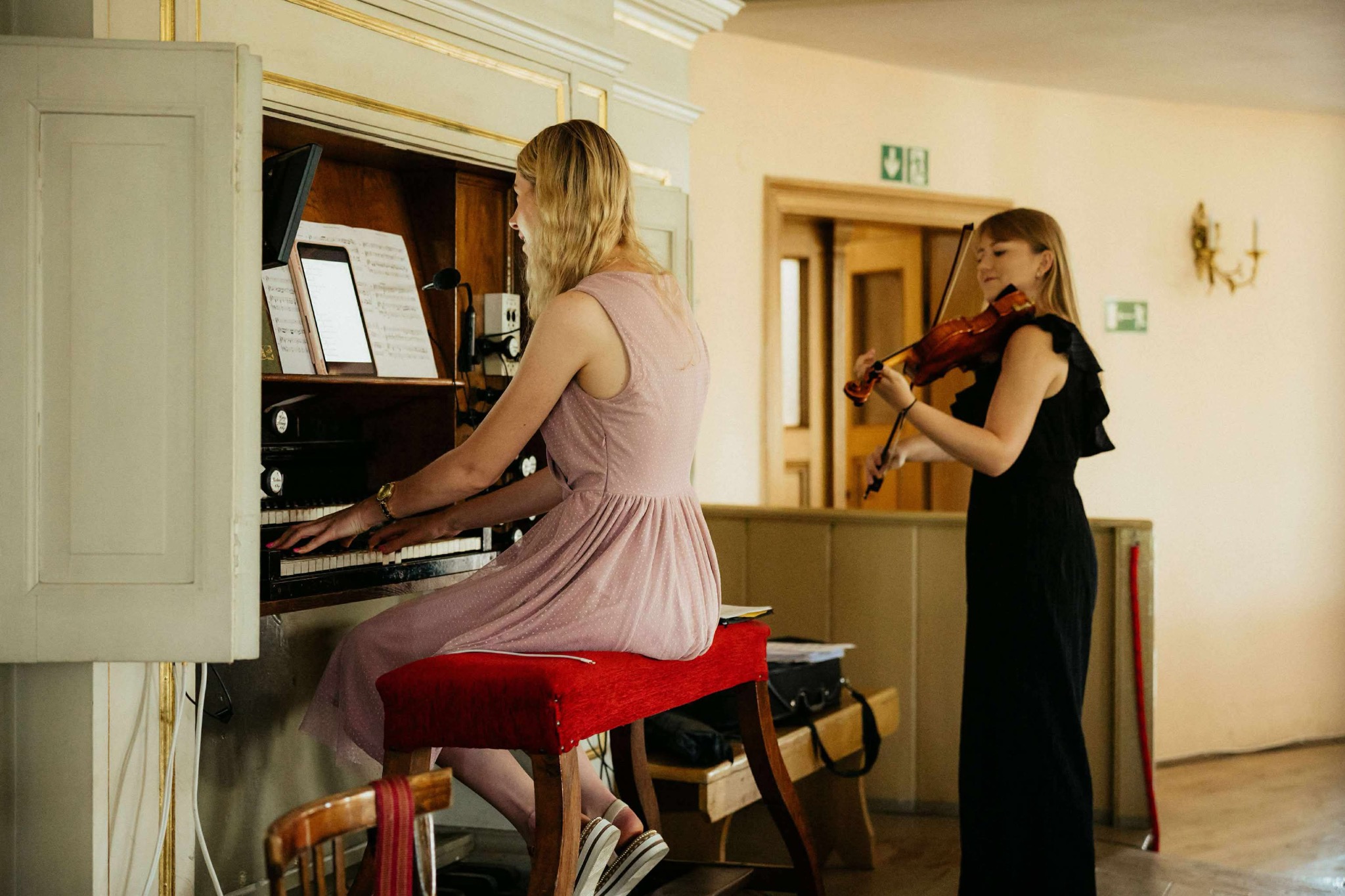 Dwie kobiety grające muzykę: jedna na organach, druga na skrzypcach. W tle jasne wnętrze z elementami dekoracyjnymi. Scena z występu muzycznego.