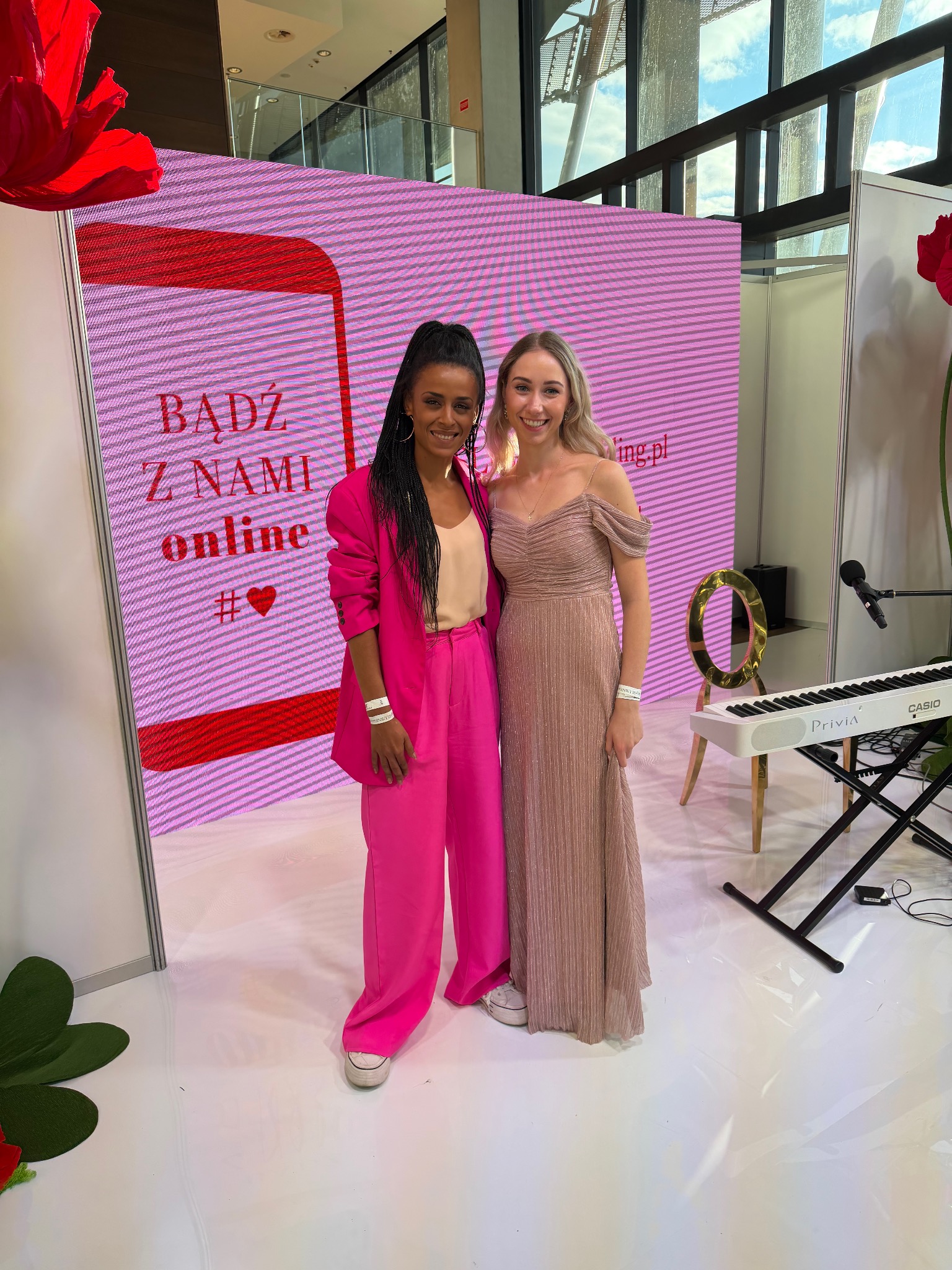 Dwie uśmiechnięte kobiety w eleganckich strojach pozują na tle sceny z pianinem Casio Privia i napisem 'Bądź z nami online'.
