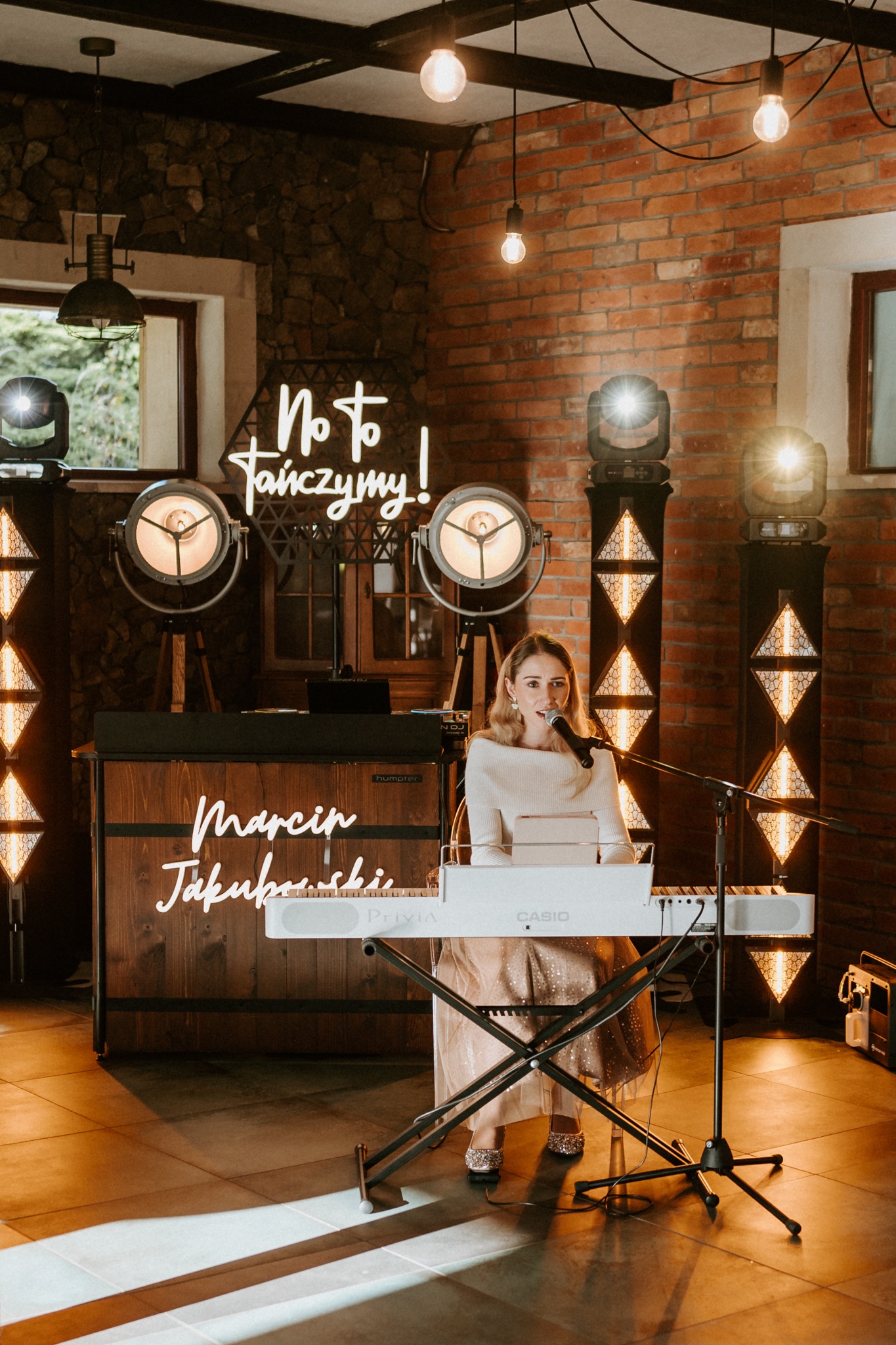 Wokalistka z mikrofonem śpiewa, grając na keyboardzie Casio. W tle ceglana ściana, neon 'No to tańczymy!' i oświetlenie sceniczne. Marcin Jakubowski.