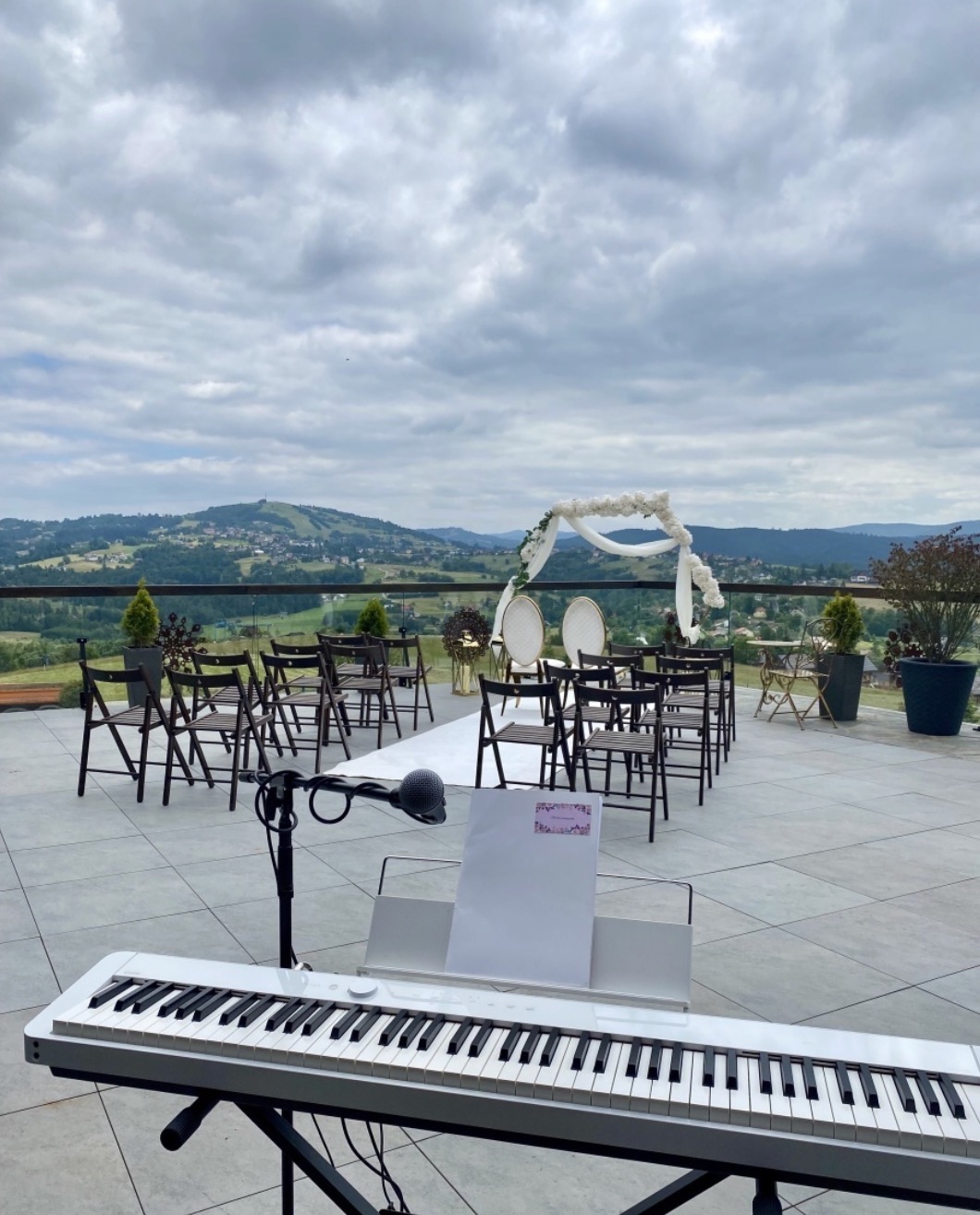 Plenerowa scena ślubna z widokiem na góry. Biały dywan, krzesła dla gości, dekoracyjny łuk i keyboard z mikrofonem na pierwszym planie. Pochmurne niebo w tle.
