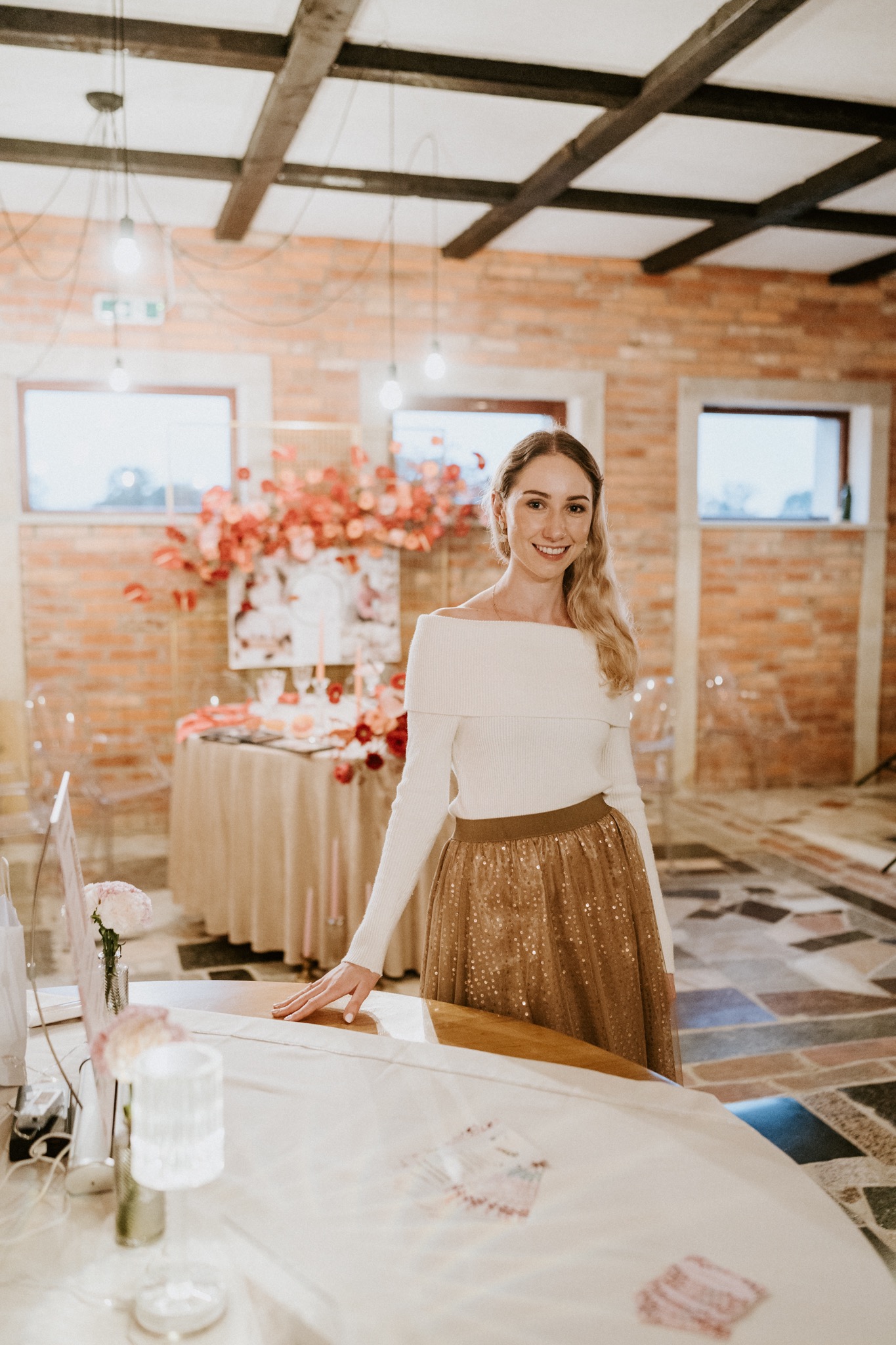 Uśmiechnięta kobieta w białym swetrze i złotej spódnicy stoi w sali z dekoracjami kwiatowymi i stołami, prawdopodobnie podczas przygotowań do uroczystości.