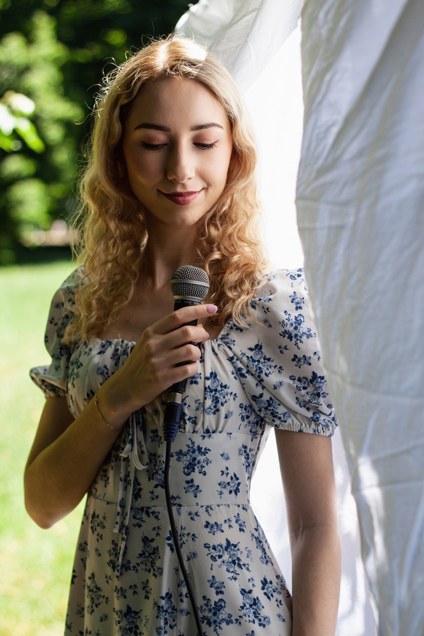 Uśmiechnięta blondynka w kwiecistej sukience śpiewa do mikrofonu Shure SM58 na tle zieleni i białej tkaniny. Delikatny makijaż, naturalne światło.