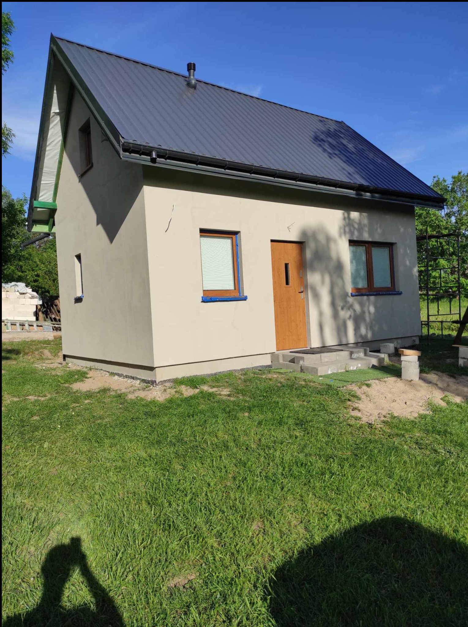 Niewielki dom z szarą blachodachówką i beżową elewacją, częściowo wykończony, z drewnianymi oknami i drzwiami. Widoczne materiały budowlane w tle.