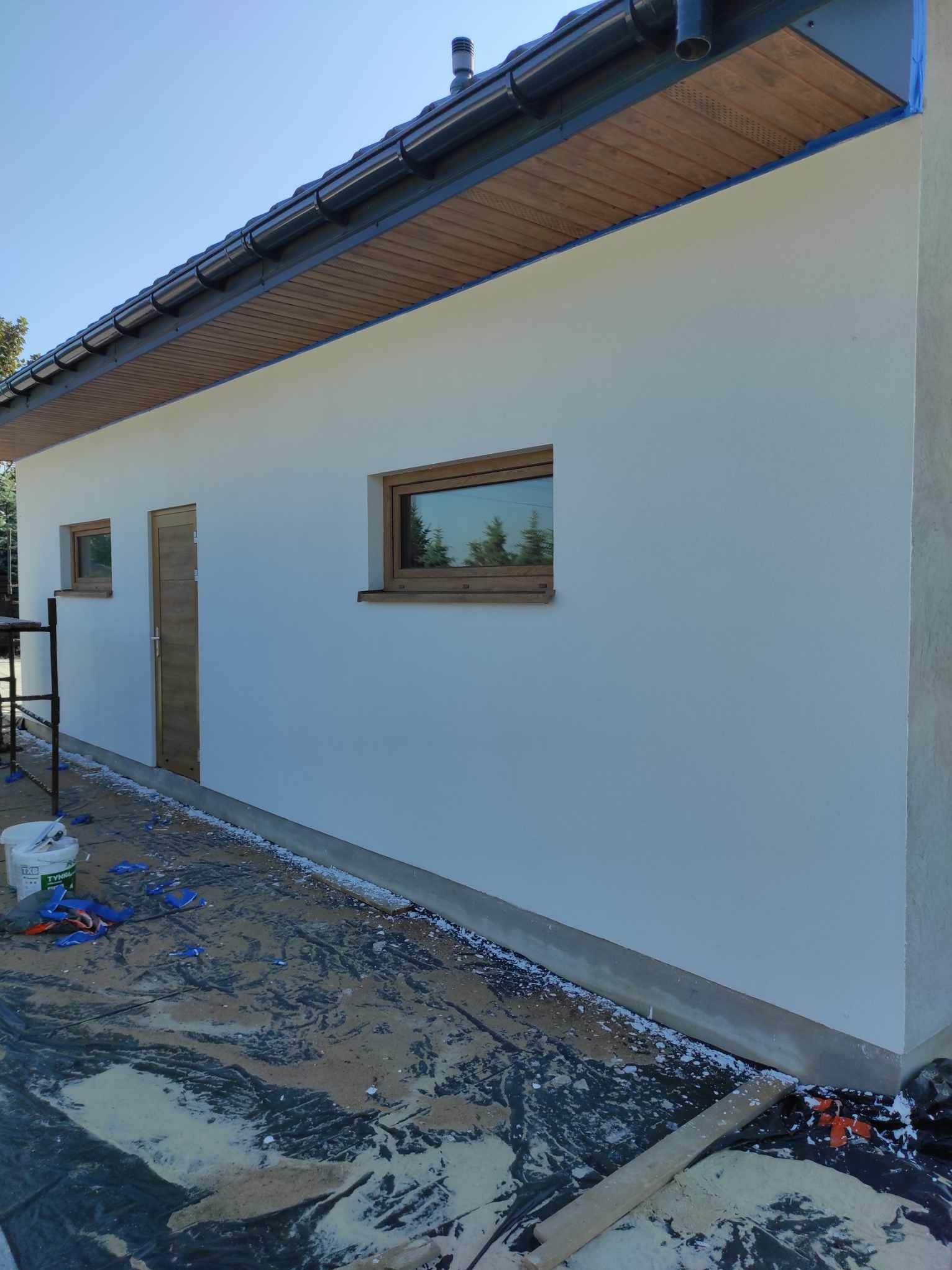 Boczny widok otynkowanego budynku z drewnianą stolarką okienną i drzwiami. Na ziemi widoczne ślady prac wykończeniowych i wiadro z farbą. Elewacja w jasnym odcieniu.