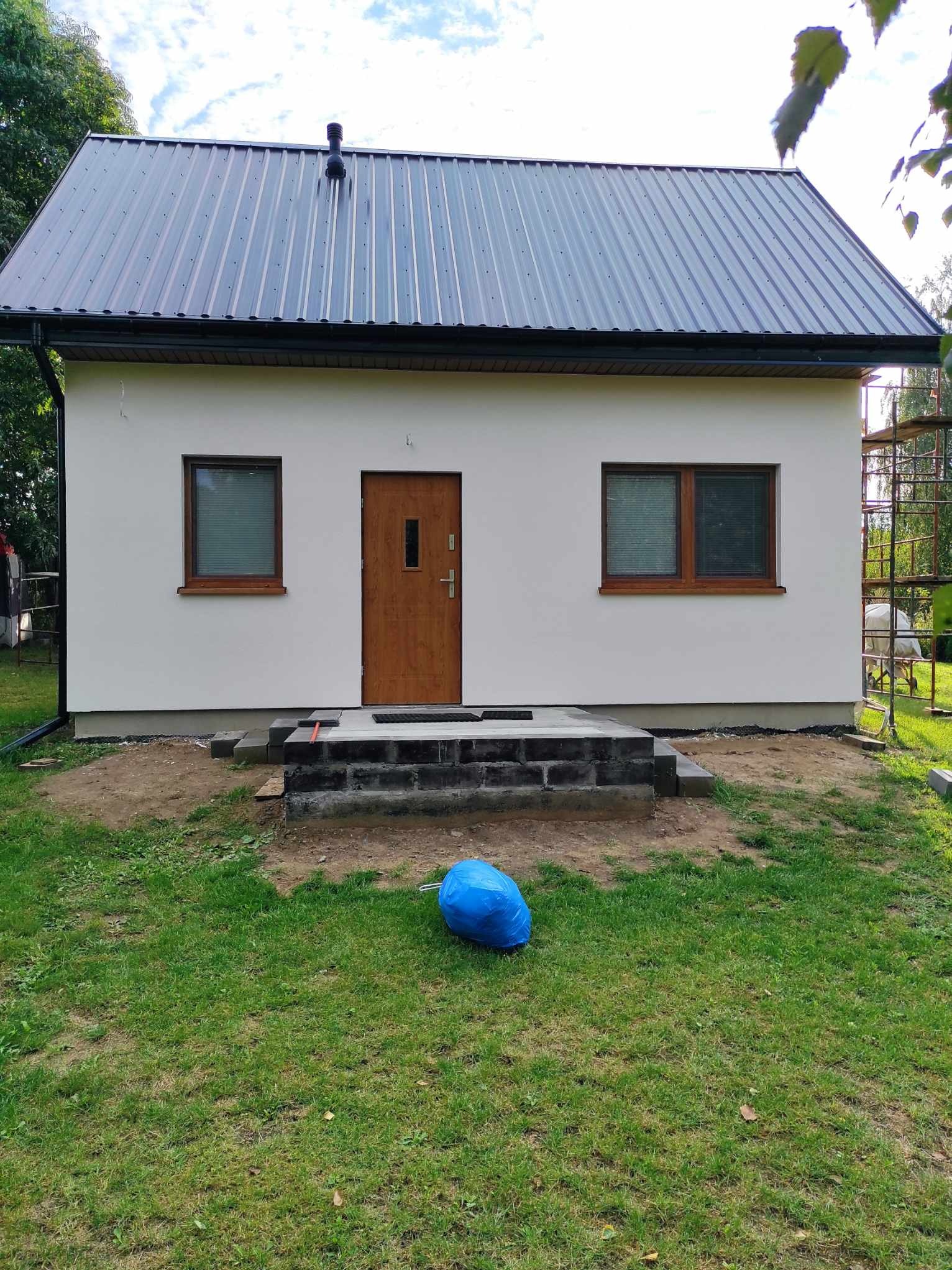 Biały, parterowy dom z ciemnym dachem z blachy trapezowej i drewnianymi oknami. Widoczne schody wejściowe w trakcie budowy i niebieski worek na trawniku.