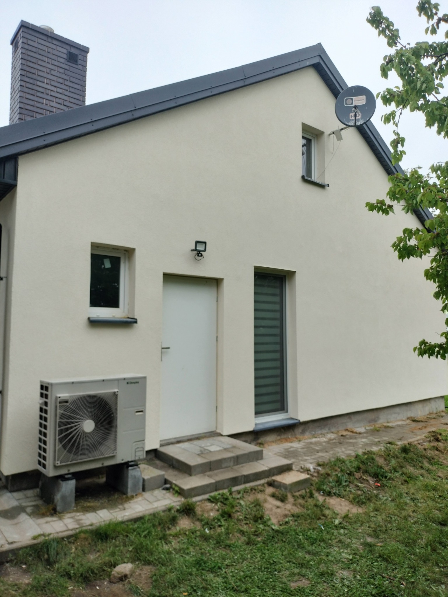 Jasna elewacja domu jednorodzinnego z klimatyzatorem, widok z boku. Białe okna i drzwi, szara dachówka i antena satelitarna. Ujęcie w pochmurny dzień.