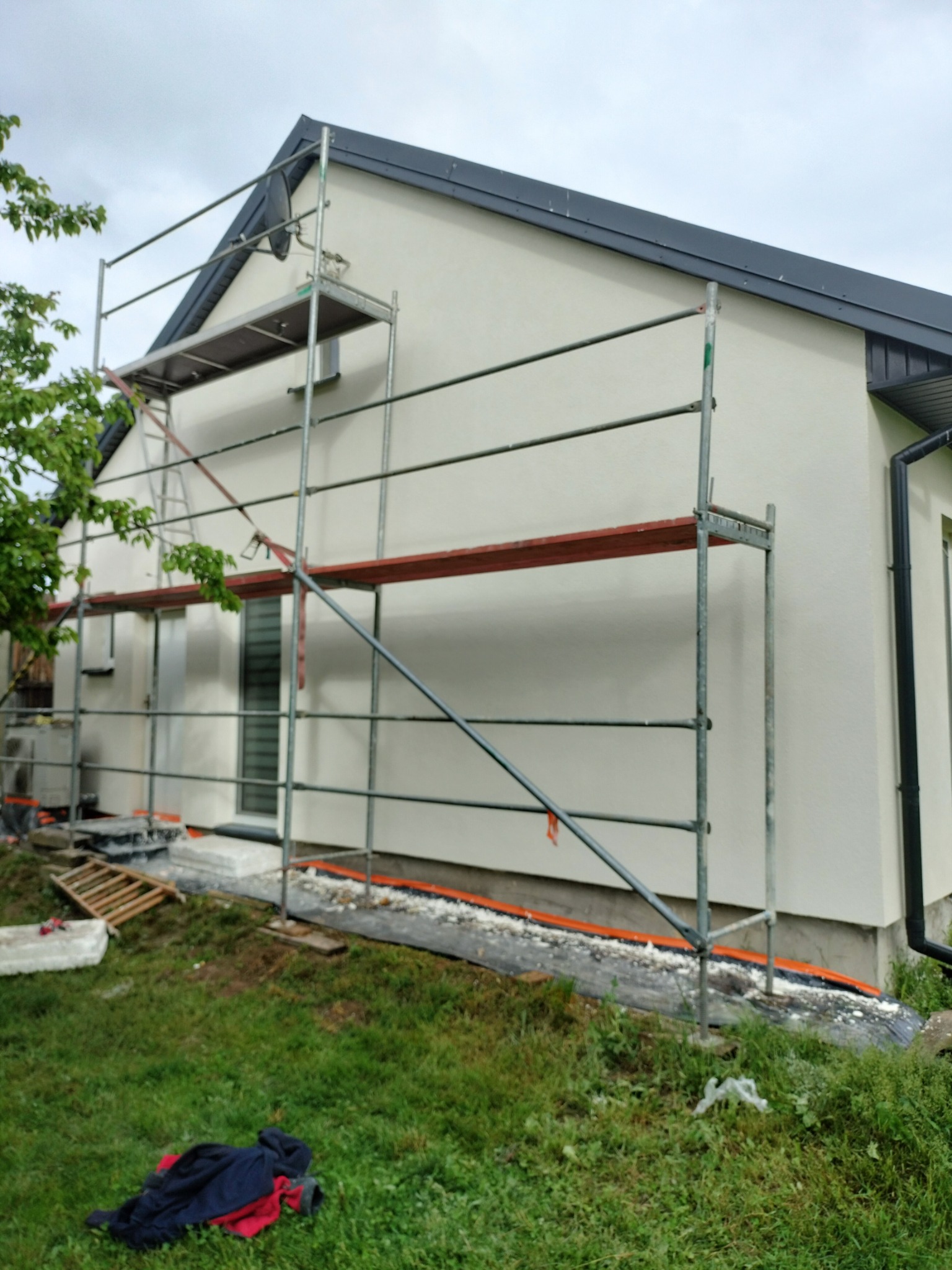 Dom jednorodzinny w trakcie prac elewacyjnych. Widoczne rusztowanie, ocieplenie przy gruncie i świeżo pomalowana ściana w jasnym odcieniu. Częściowo zachmurzone niebo.